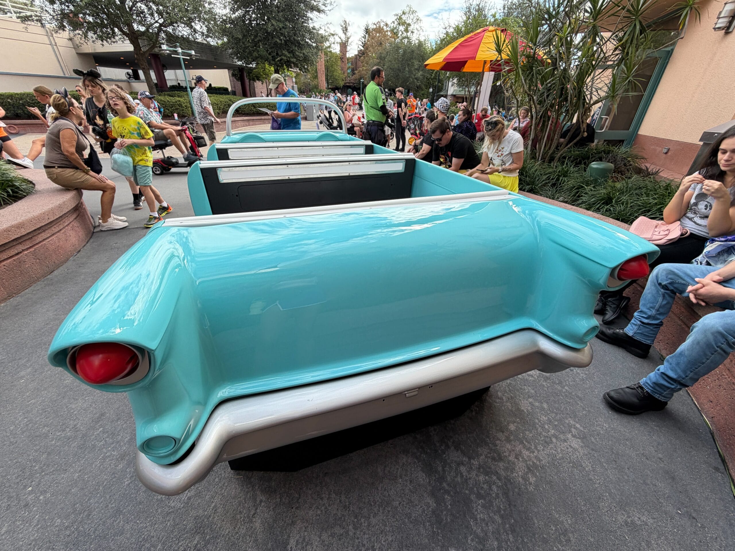 A blue vintage car-shaped bench with people seated around it in a sunny Disney park area, trees and umbrellas nearby.
