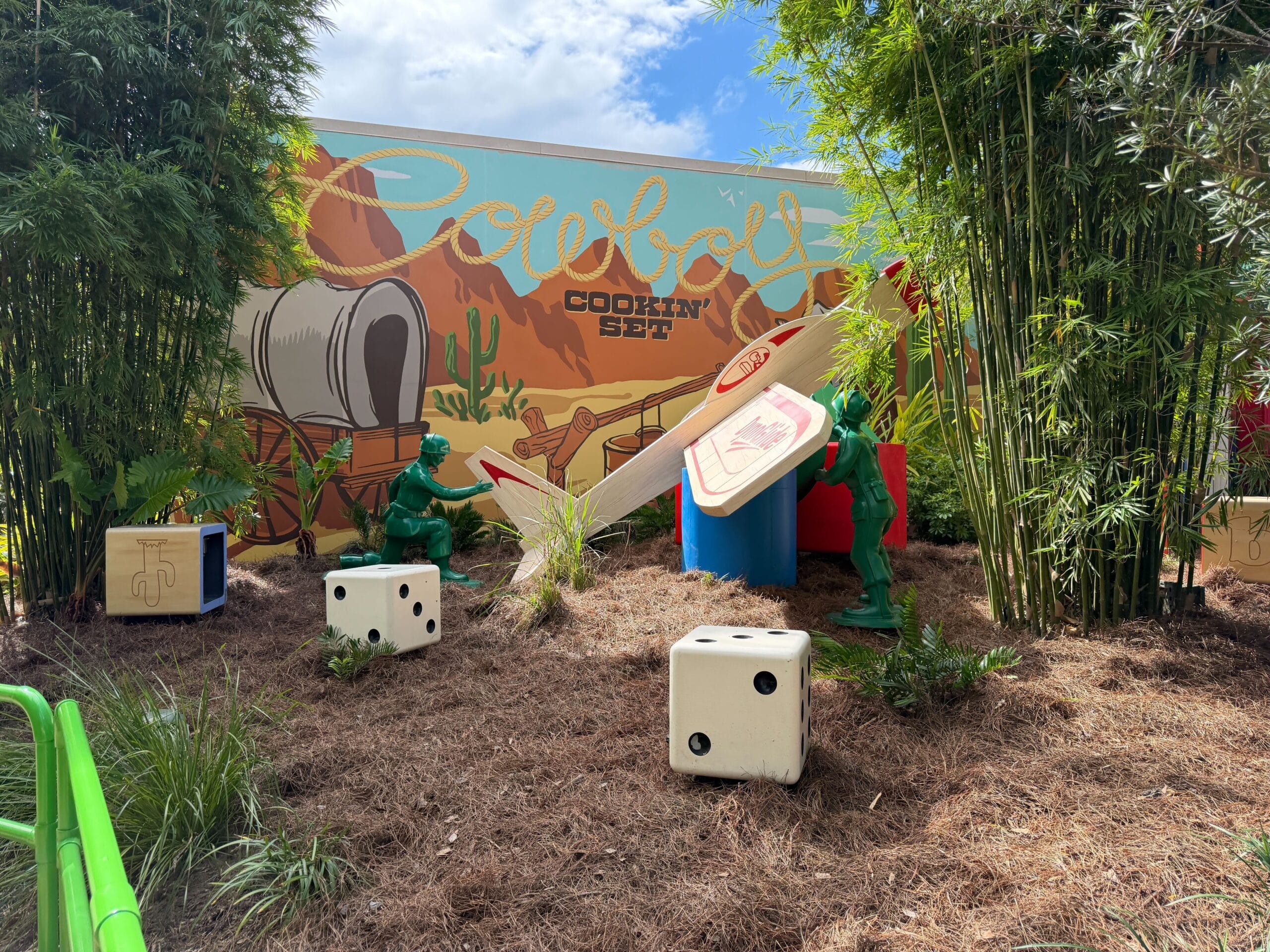 Toy soldiers play with giant dice and a cracker in a garden, resembling Pixar Play Parade areas at Disneyland, western mural behind.