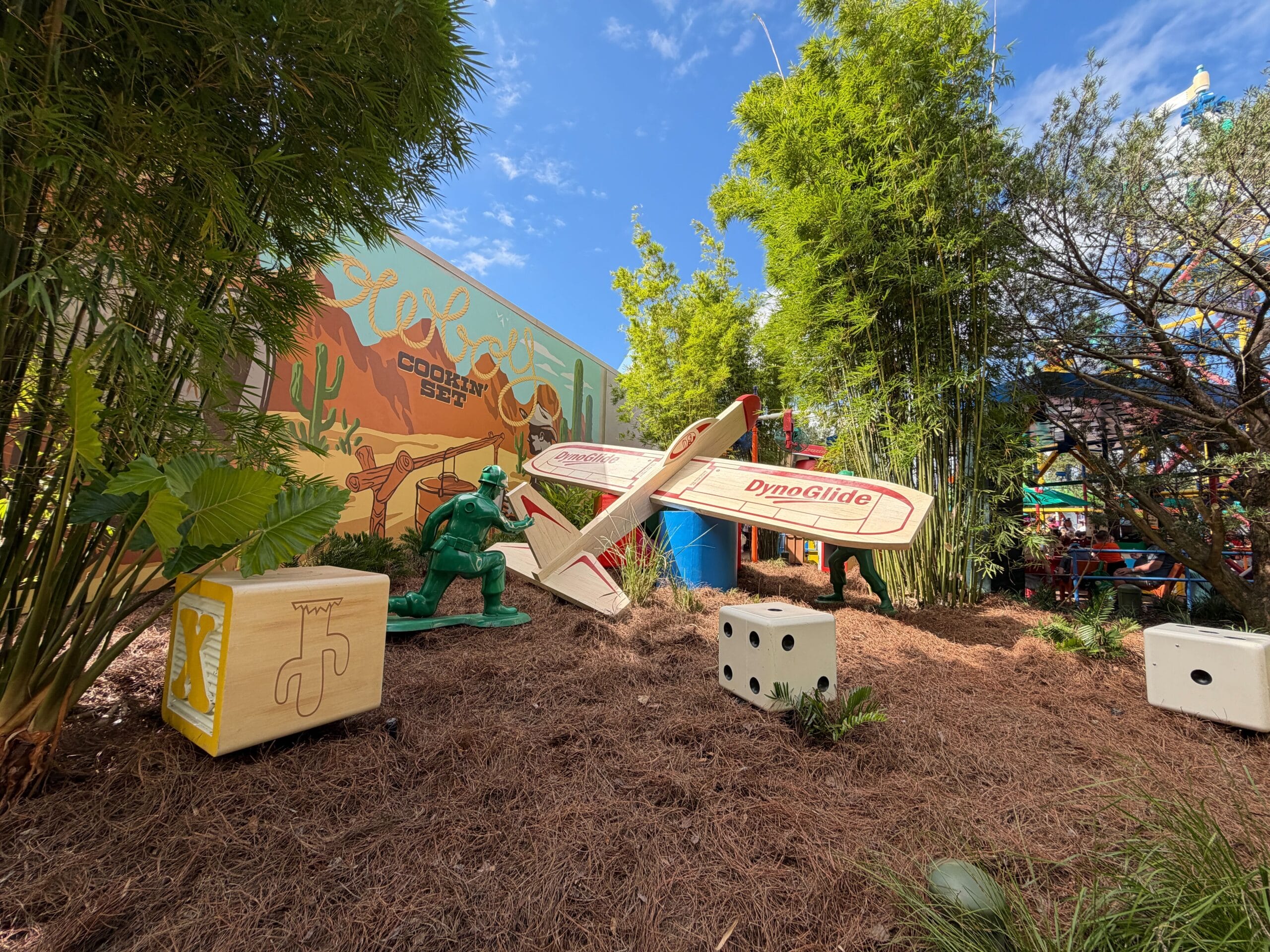 A toy plane, oversized dice, and a green dinosaur figure are displayed in a garden with a vibrant mural, Toy Story Land style.