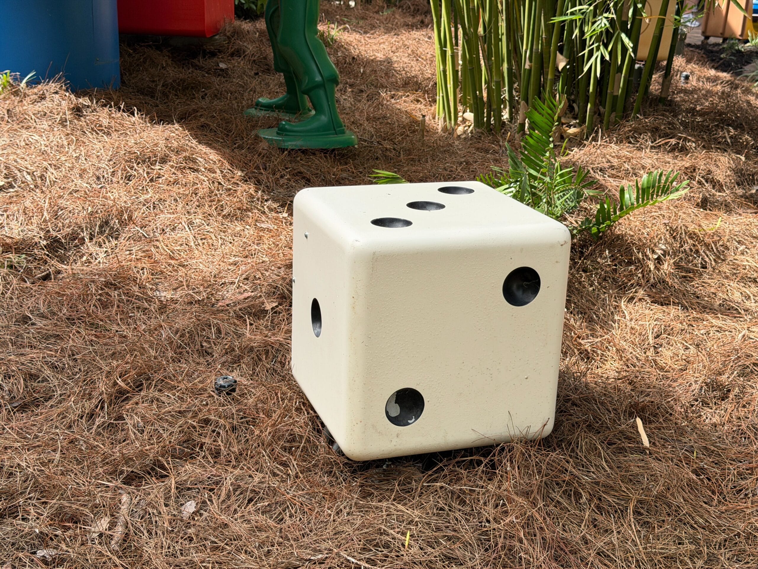 A large white die with black dots rests on pine needles near plants and a green toy, reminiscent of Toy Story Land in Disney parks.