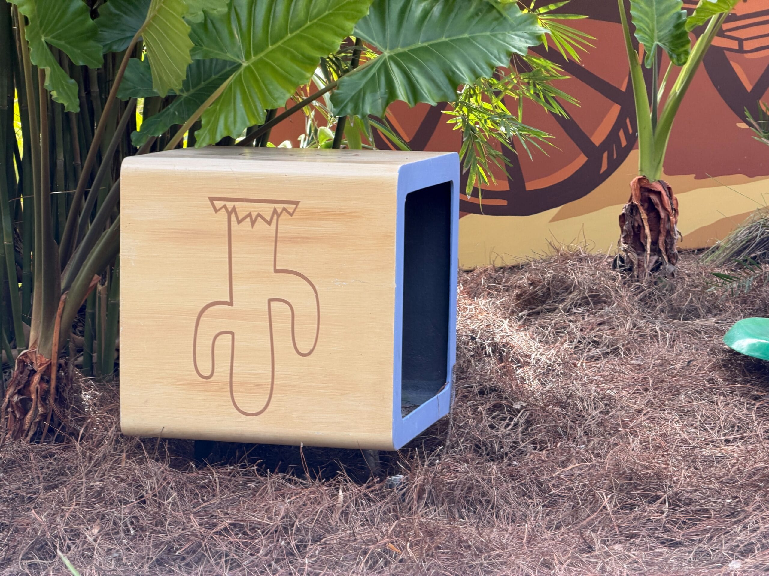 A wooden box with a carved plant design rests on pine straw by lush tropical foliage, similar to Adventureland in Disney parks.