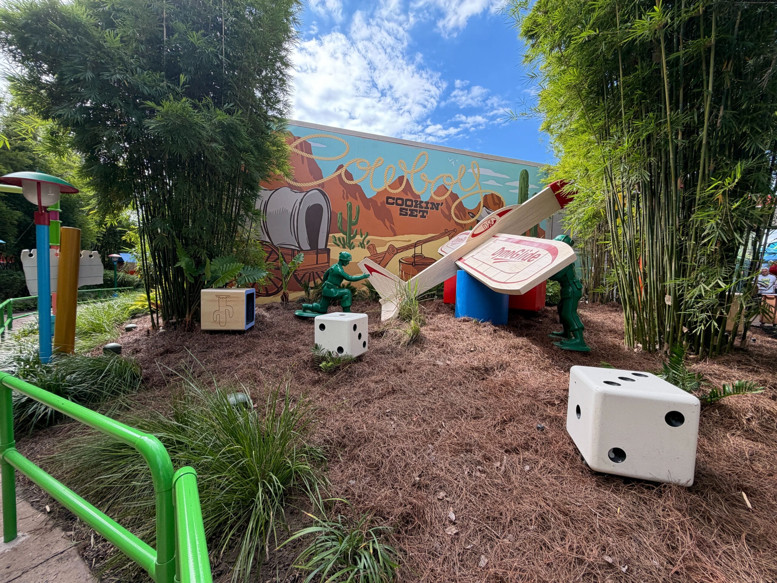 Toy soldiers, oversized dice, and game pieces decorate a garden with a vibrant mural and bamboo, resembling Disney park play areas.
