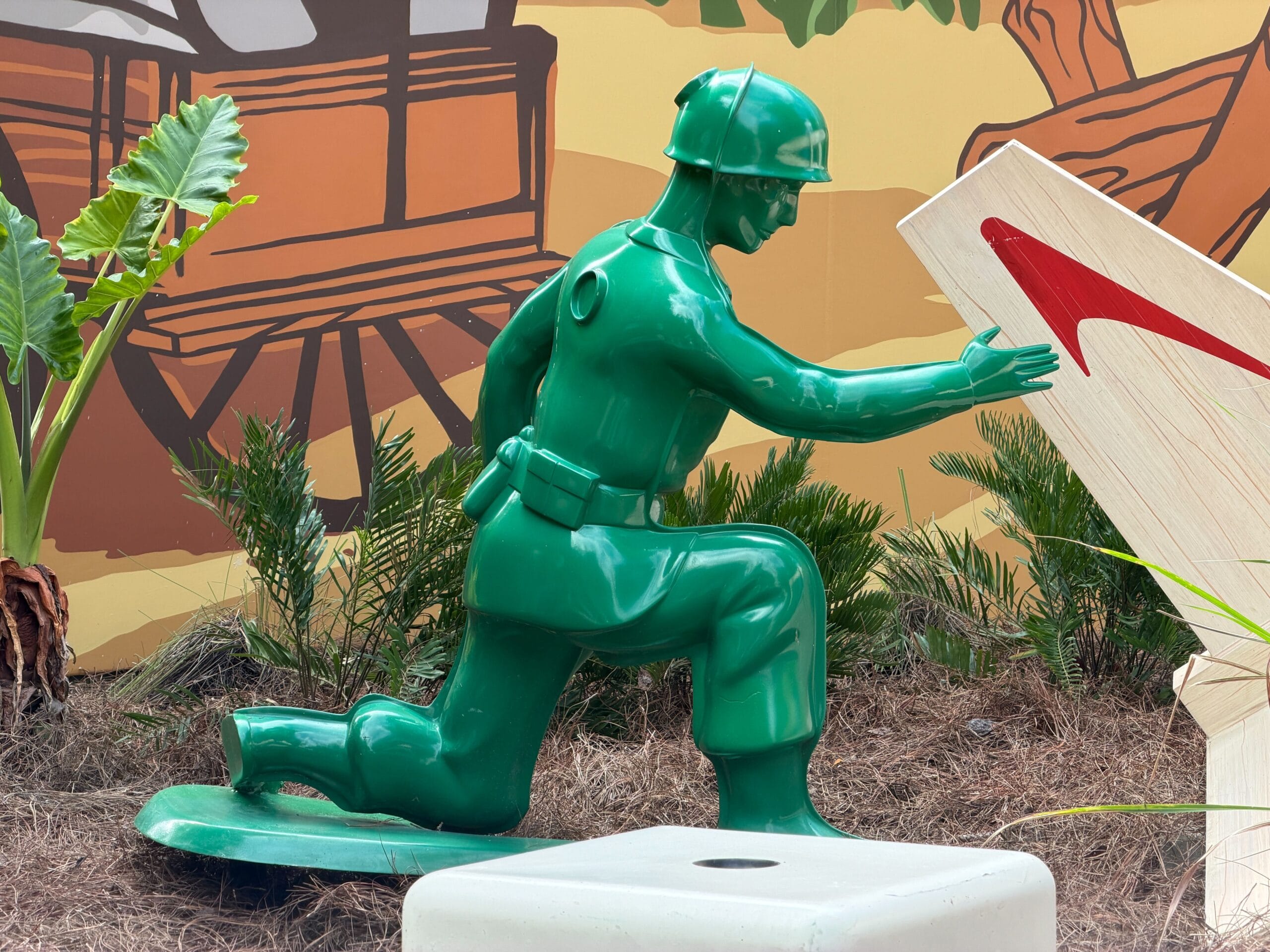 A large green toy soldier statue kneels on one knee outdoors in playful, cartoonish landscaping, reminiscent of Disney parks decor.