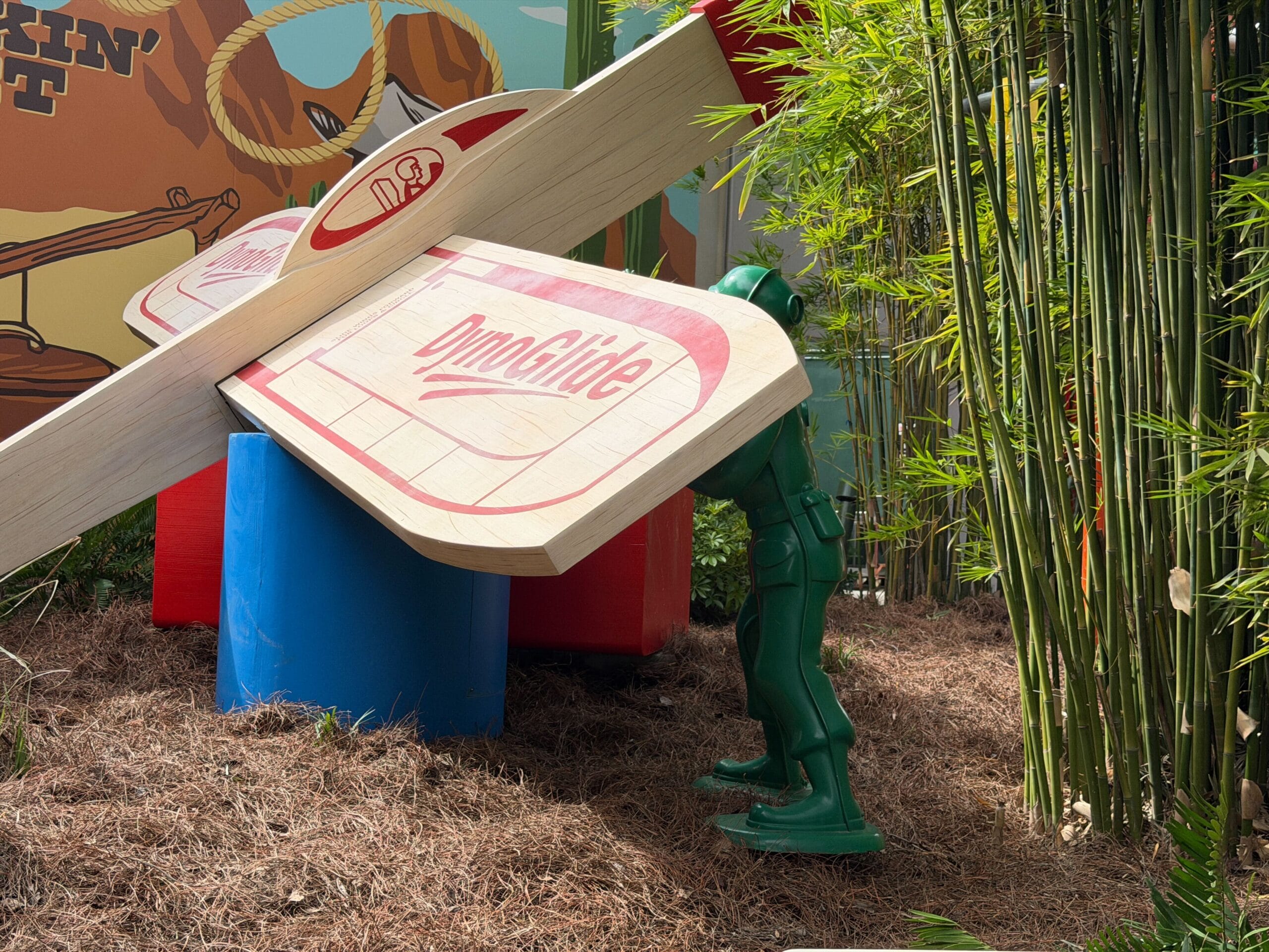 Green toy soldier with large wooden glider wing stands by bamboo and bright props, similar to Toy Story Land displays in Disney parks.