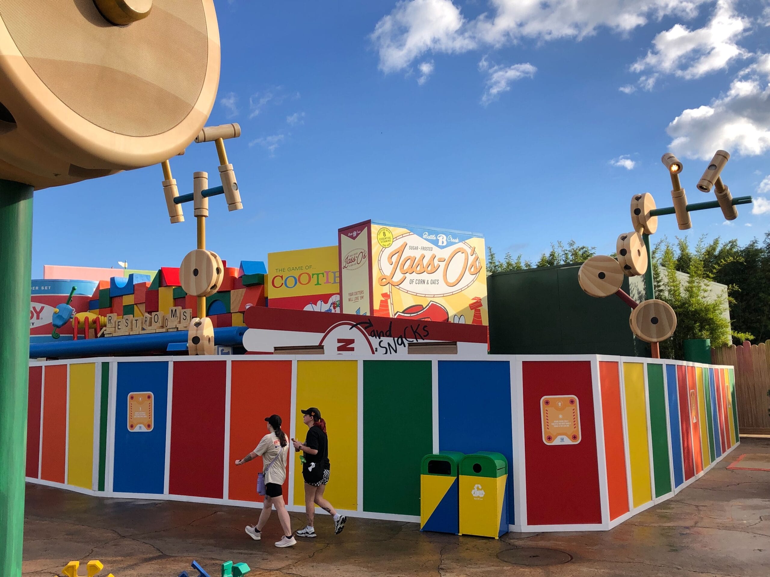 Two guests stroll by a Toy Story-themed construction wall at a Disney park, vivid sky and clouds overhead, no castle in sight.