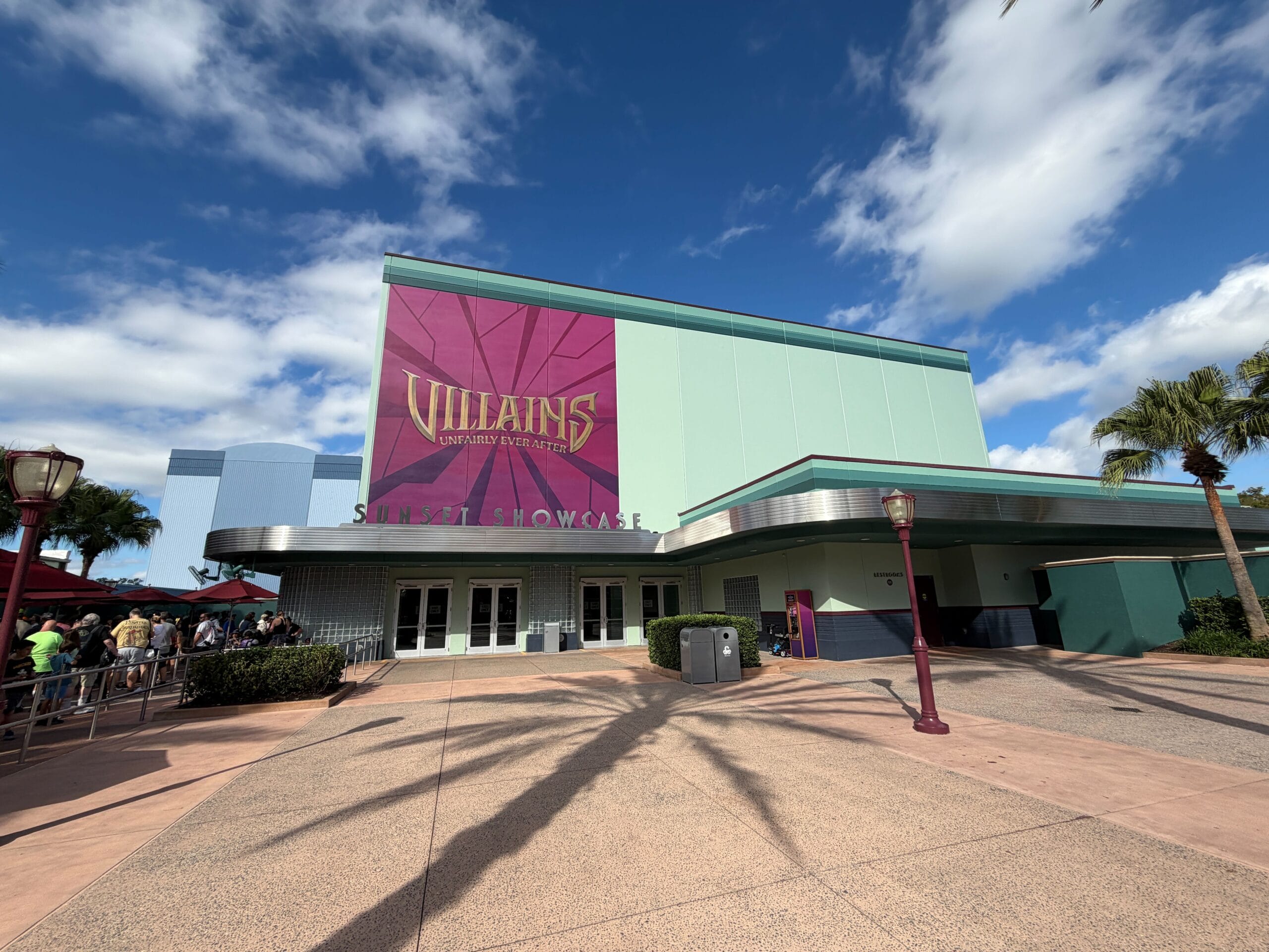 A large building with a "Villains" sign out front, reminiscent of Disney park theming, beneath a bright blue, cloud-dotted sky.