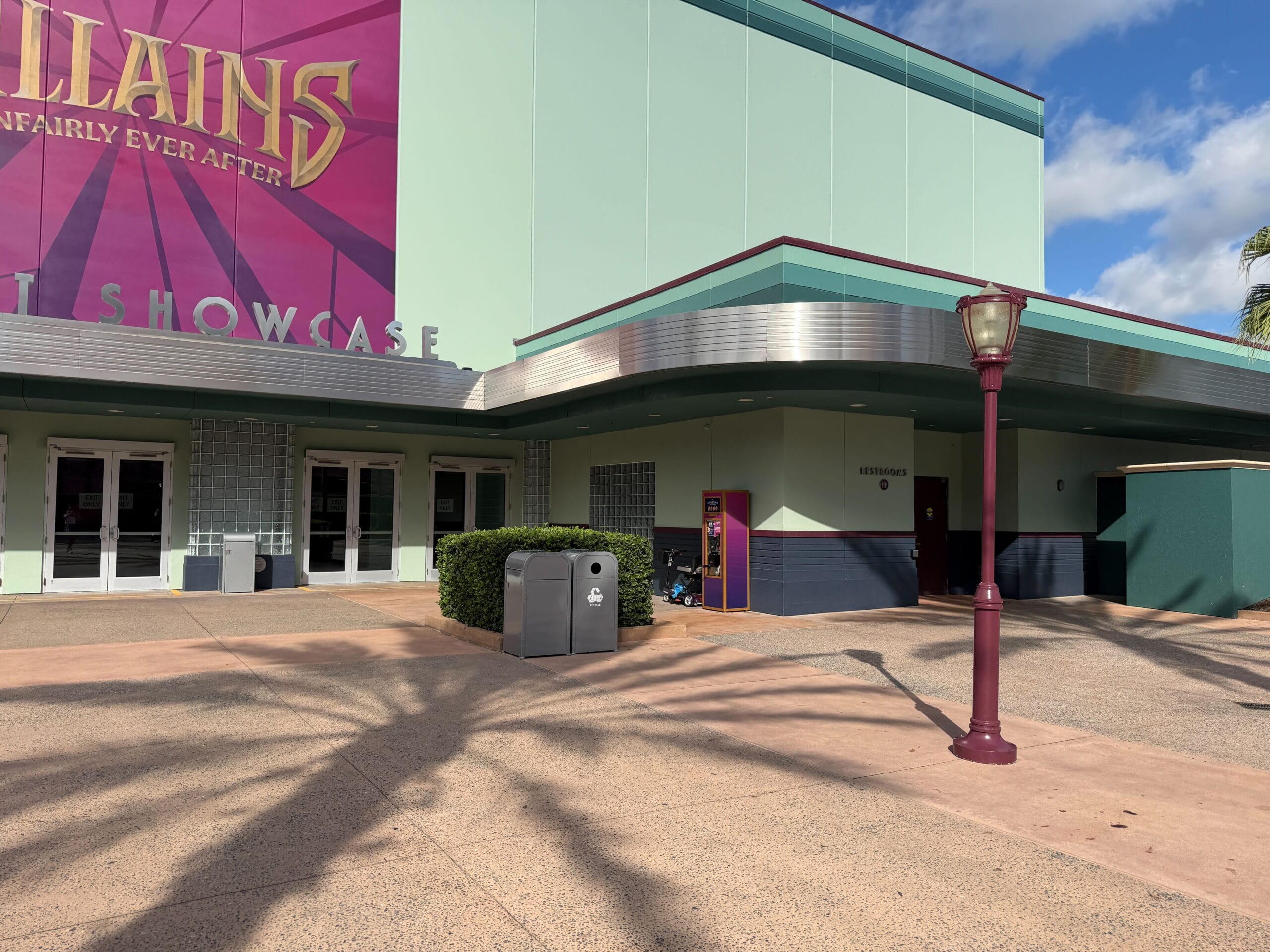 A peaceful building exterior in a Disney park, featuring a pink streetlamp, themed trash cans, and a “SHOWCASE” sign partly visible.