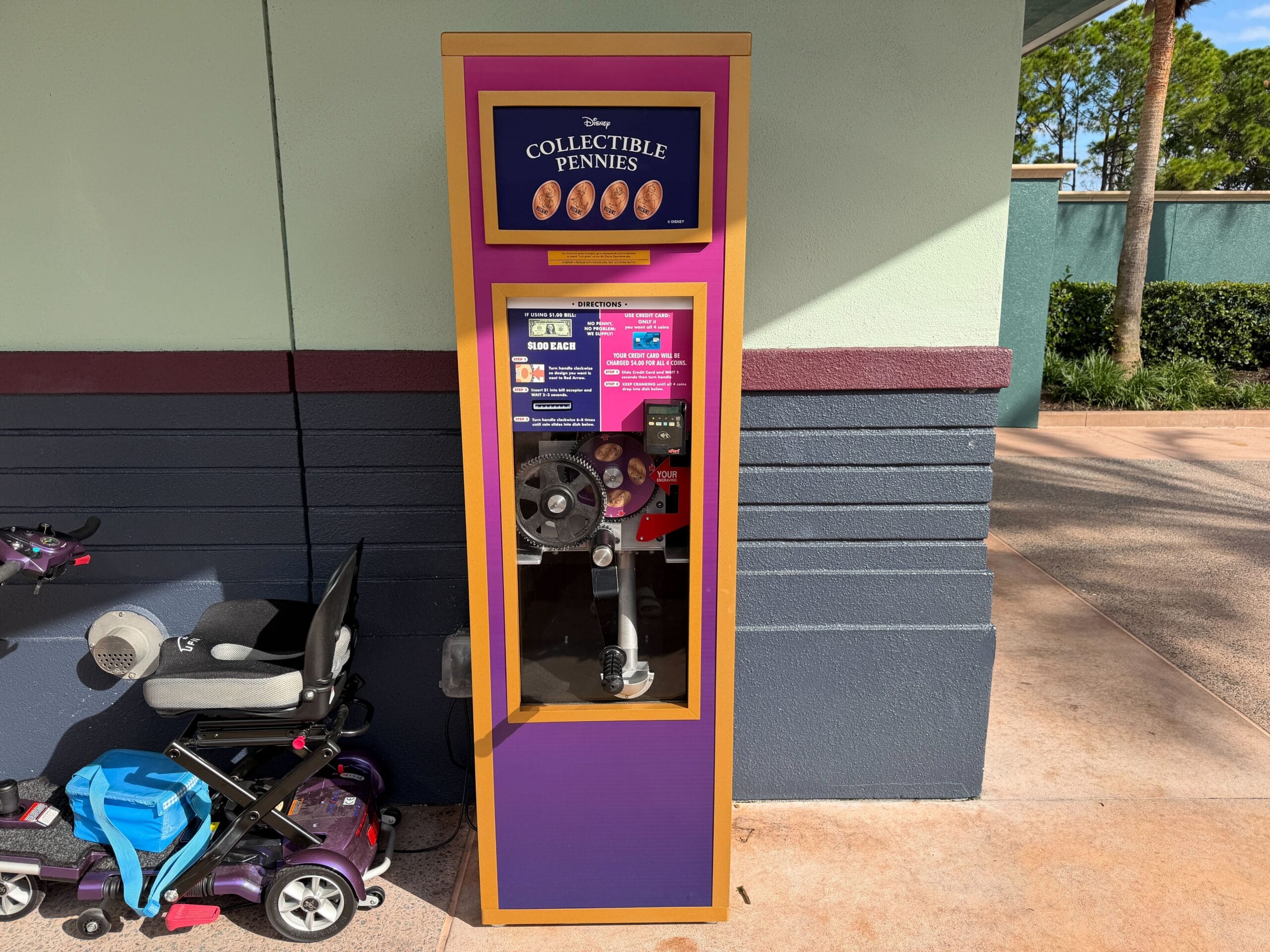 A Disney pressed penny machine stands by a wall near a parked stroller in the sun, like those commonly found across Disney parks.