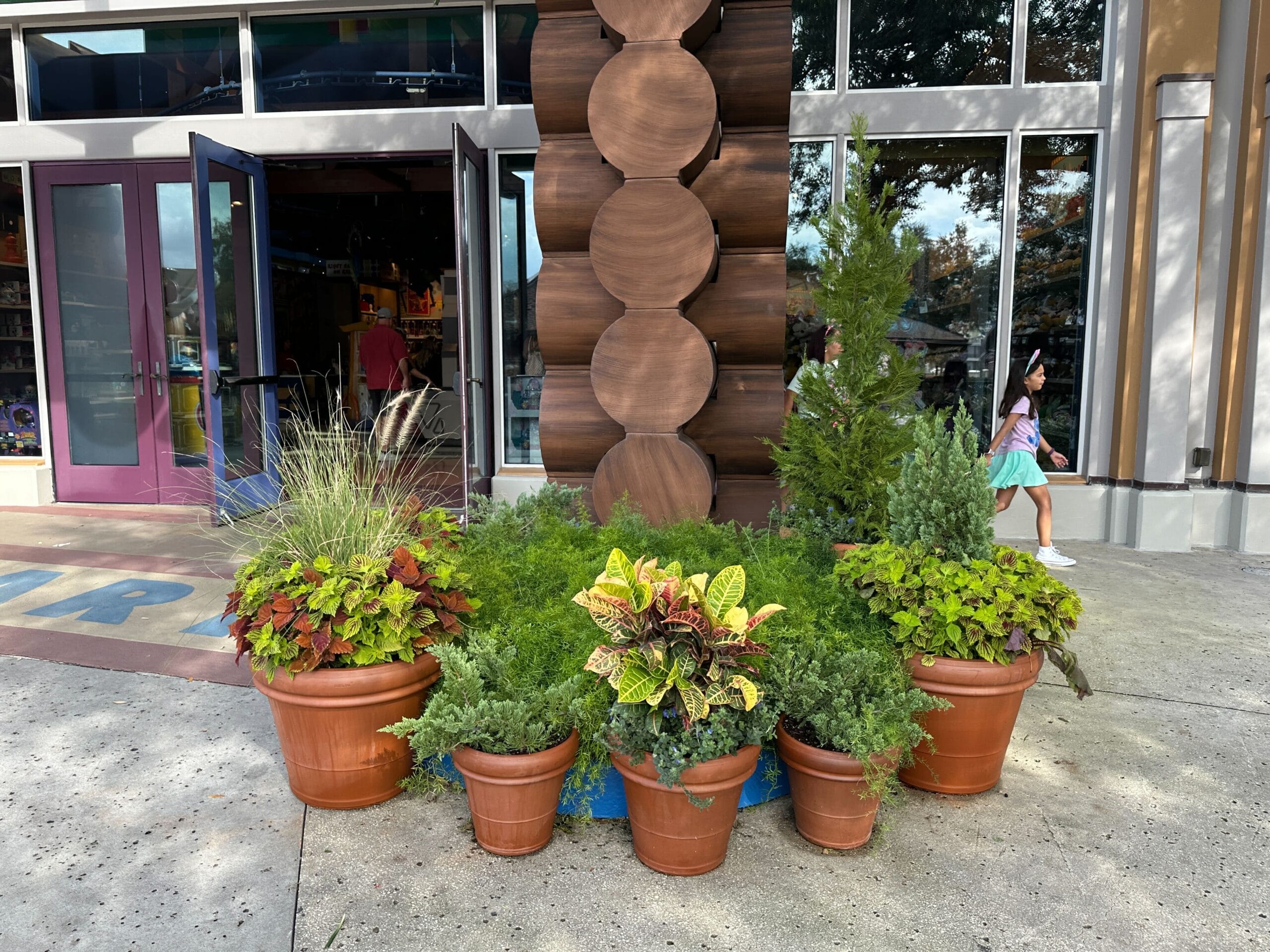 Seven potted plants line a pillar at Once Upon a Toy in Disney Springs as guests walk past the Buzz Lightyear statue nearby.