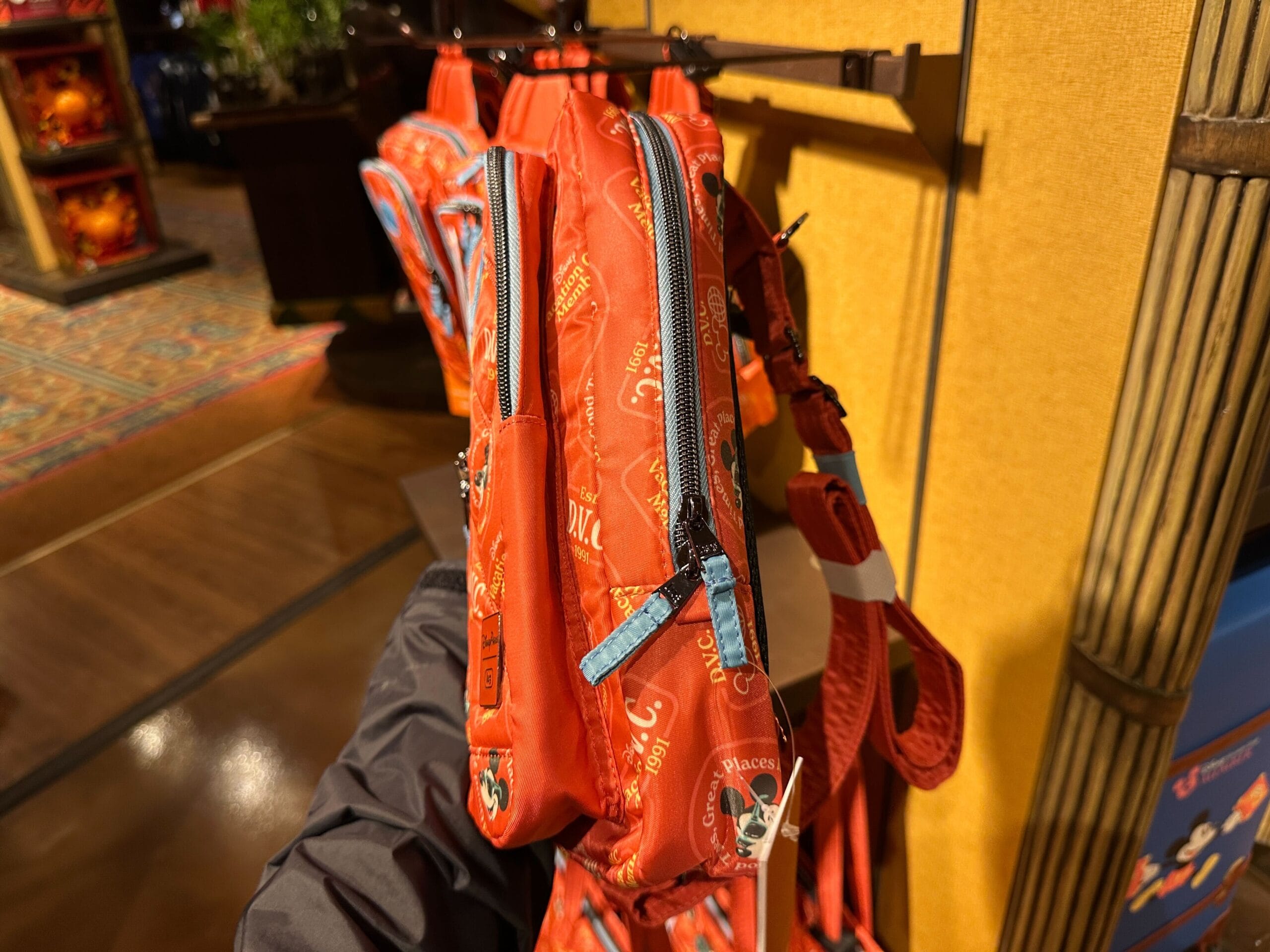 A hand holds a red backpack with Mickey designs, displayed among DVC Member Merch on a store rack.