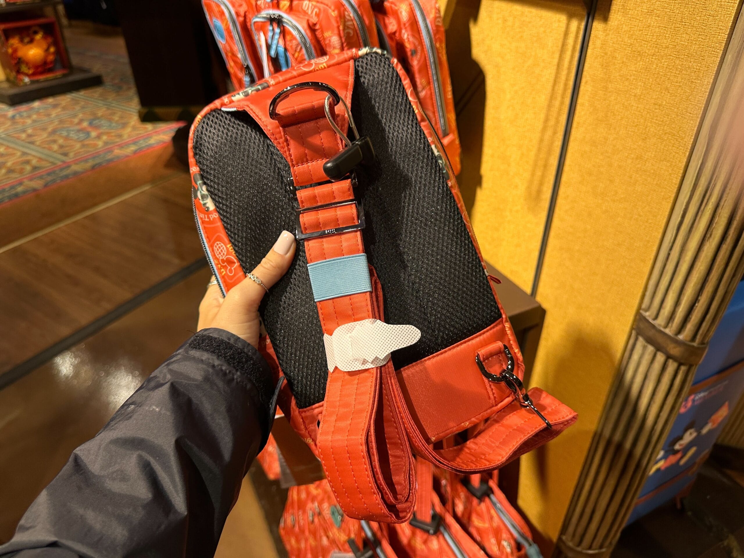 Hand holding a red and black backpack with a buckle, featuring DVC Member Merch, inside a store with more backpacks.