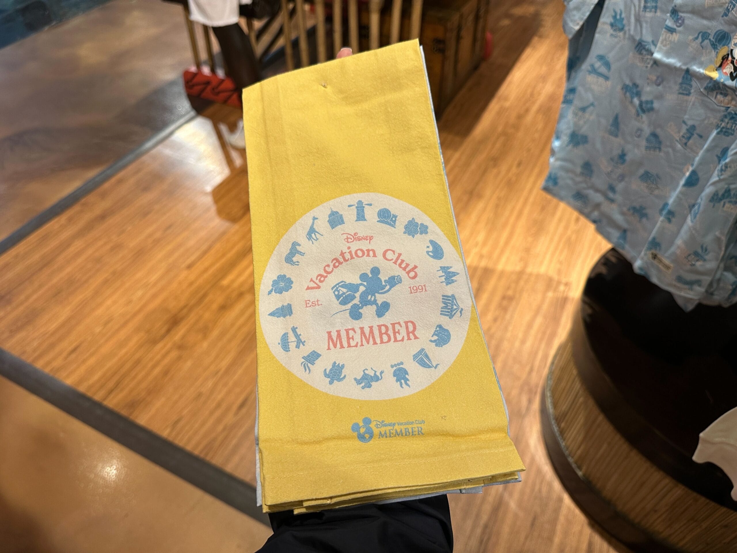 A hand holds a yellow DVC Member Merch napkin with blue icons at Disney's Grand Floridian Resort store.