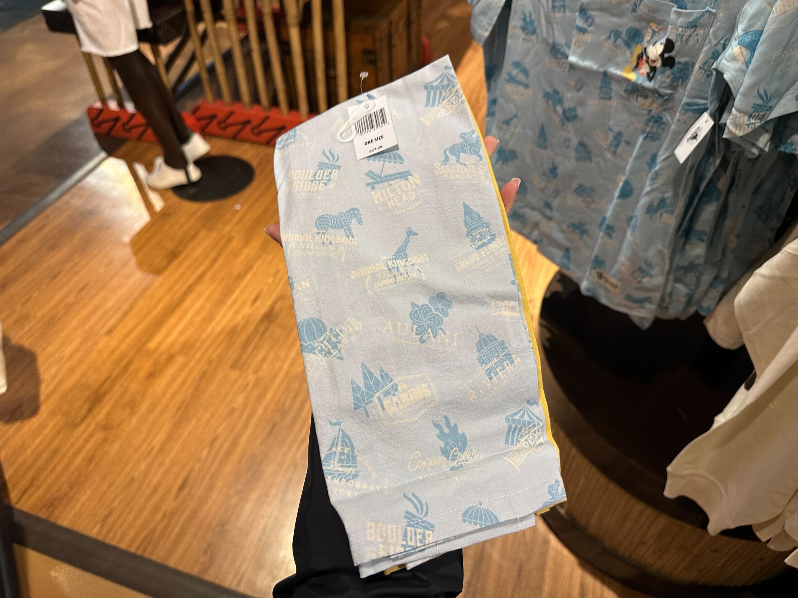 Hand holding a light blue souvenir towel with Mickey and Disney’s Grand Floridian Resort icons in a retail store.