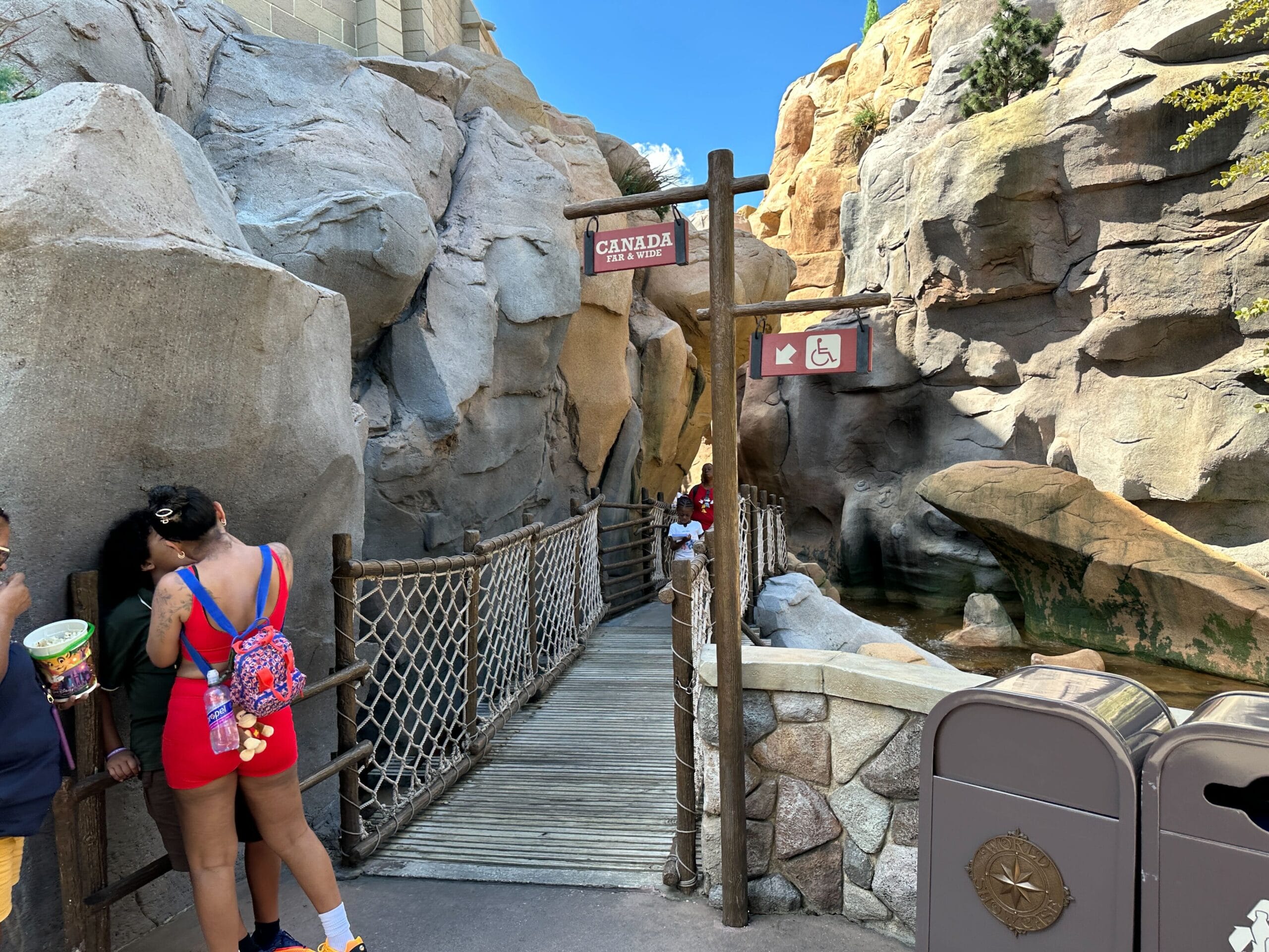 A wooden bridge between rocky cliffs in the Canada Pavilion at EPCOT, with "Canada" and accessible signs, guests nearby.