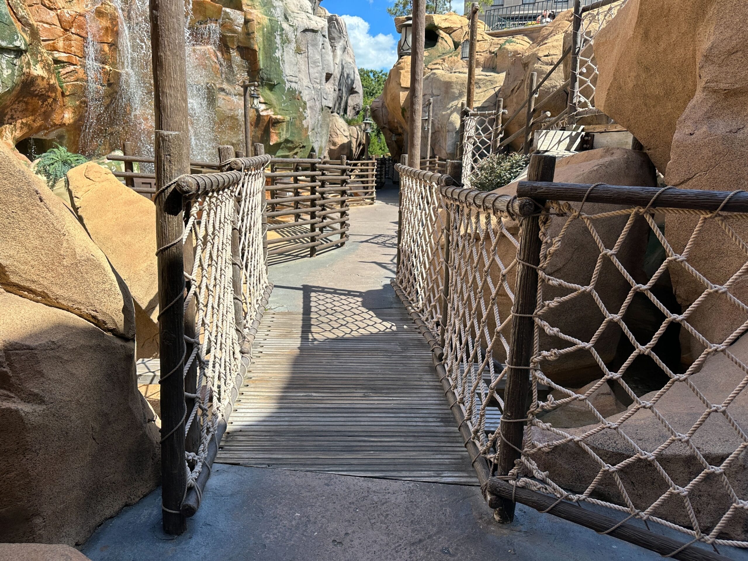 A wooden rope bridge spans rocky cliffs under a sunny sky, reminiscent of adventure bridges in Disney parks like Adventureland.