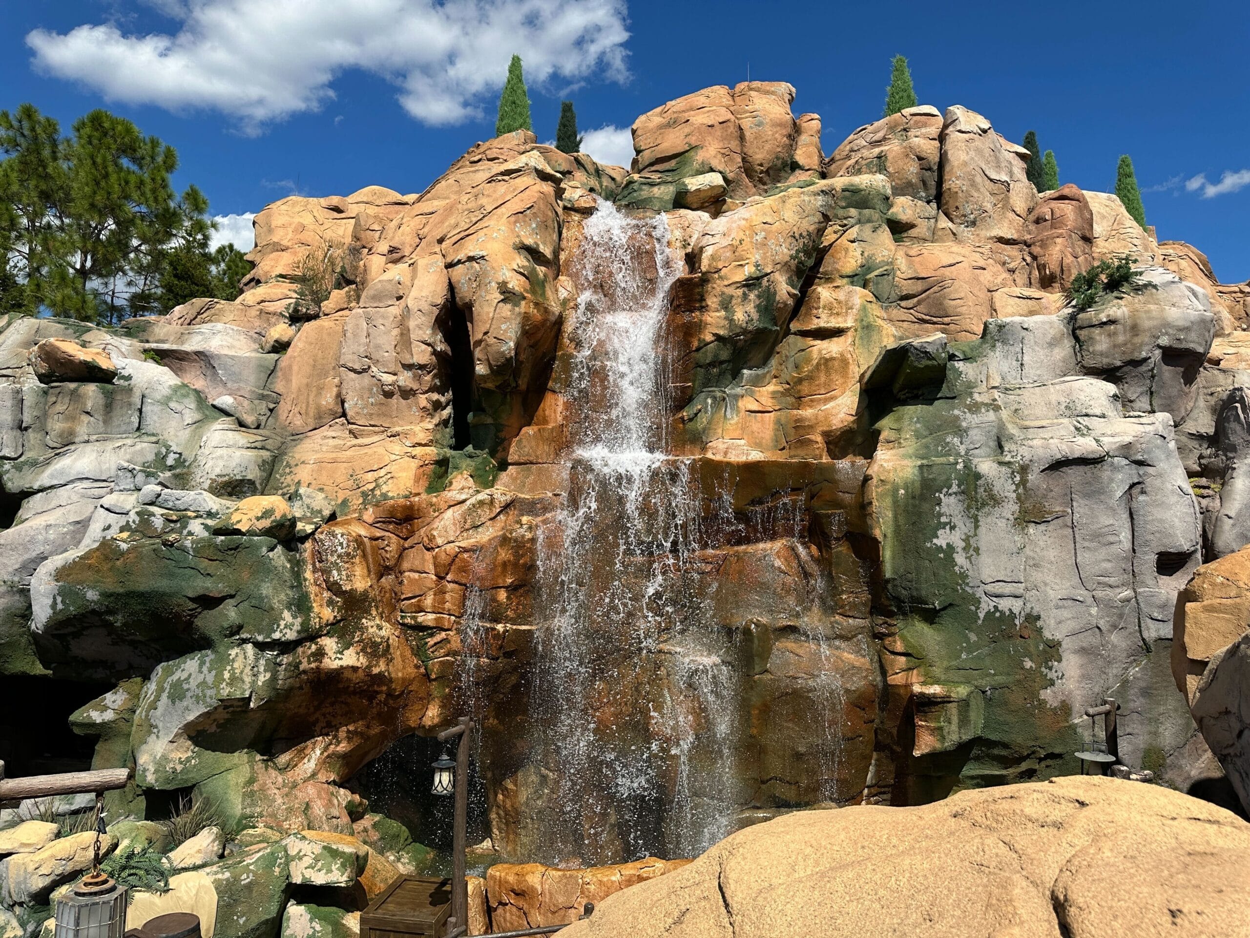 A waterfall cascades down a rocky cliff beneath a blue sky with scattered clouds, surrounded by lush greenery like in Disney parks.