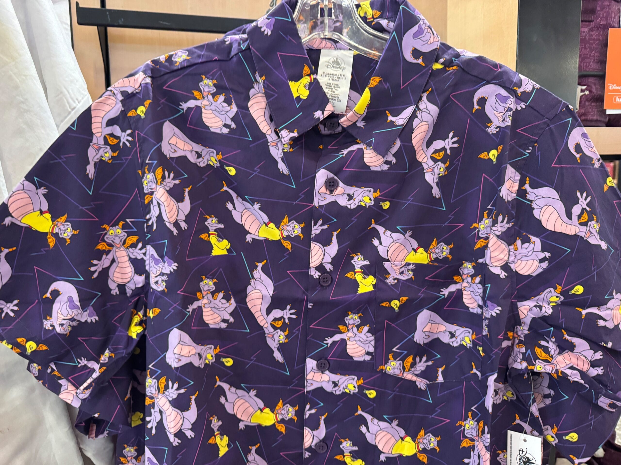 Purple button-up shirt with cartoon dragons and yellow birds, reminiscent of whimsical Disney merch, hanging in a park gift shop.