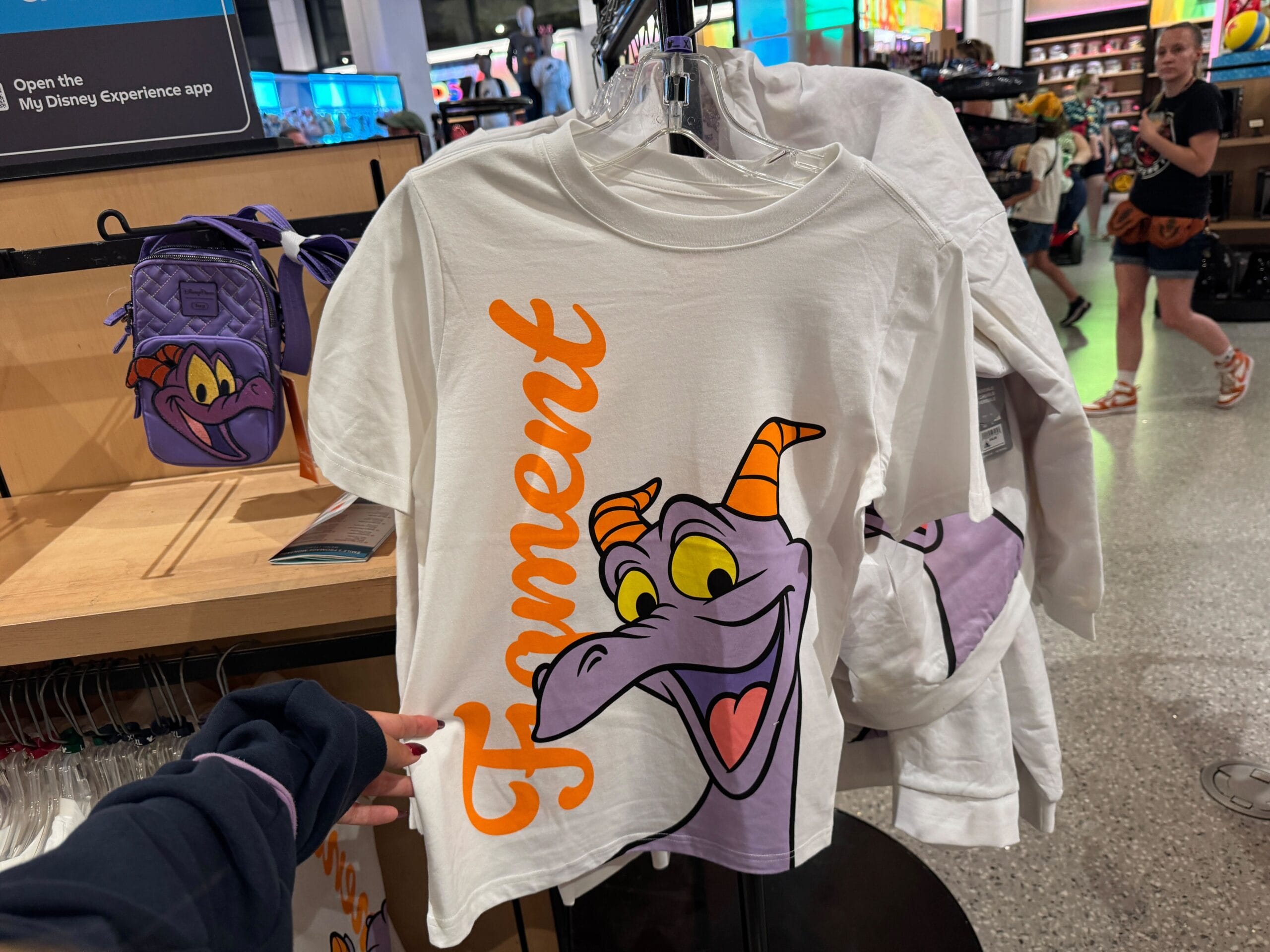 White T-shirt featuring Figment’s cartoon face and his name in orange, displayed in a Disney park store, likely Epcot.