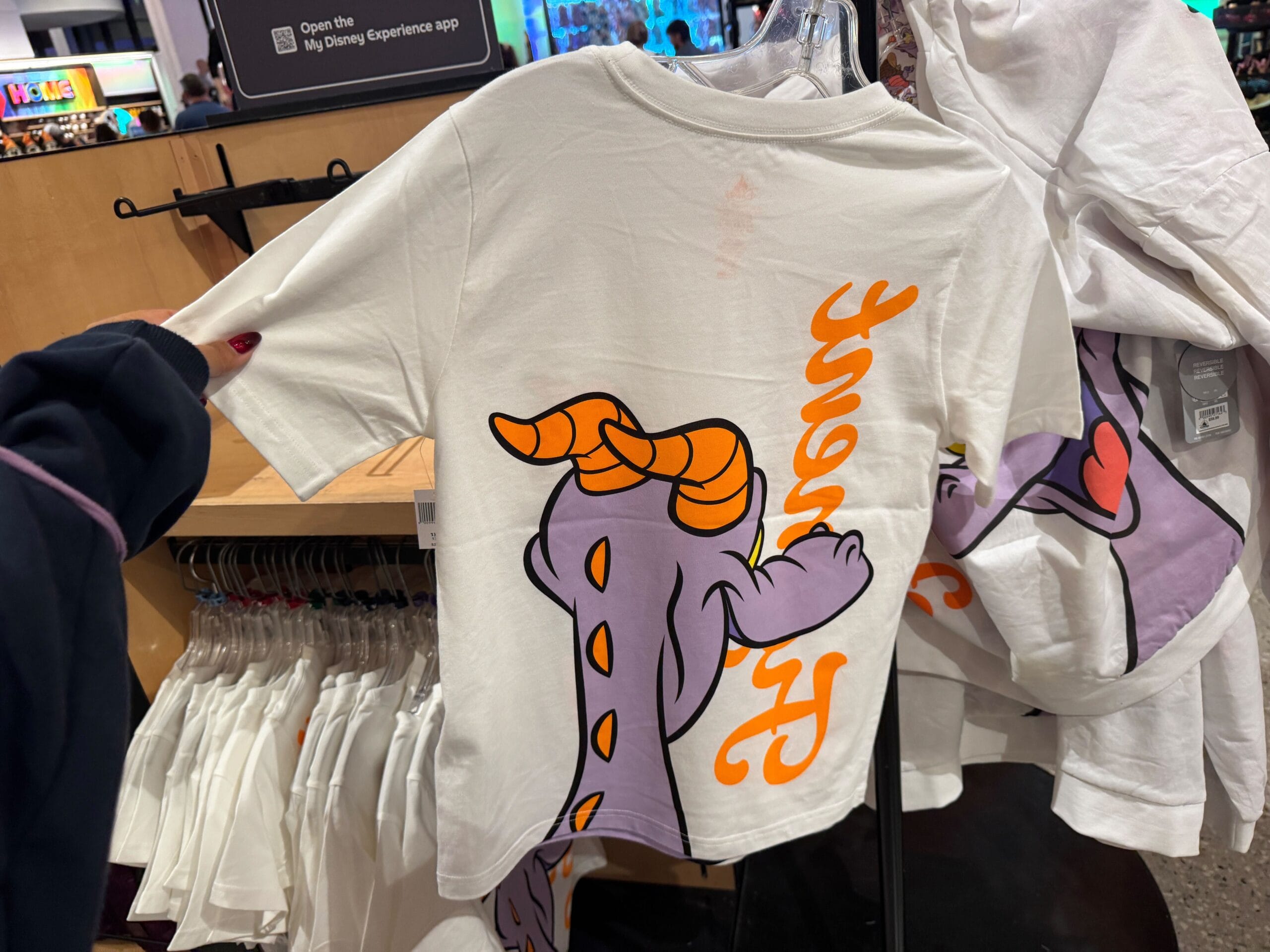 White t-shirt with a cartoon purple dragon, reminiscent of Disney’s Figment, and orange text, seen on a store rack at a Disney park.