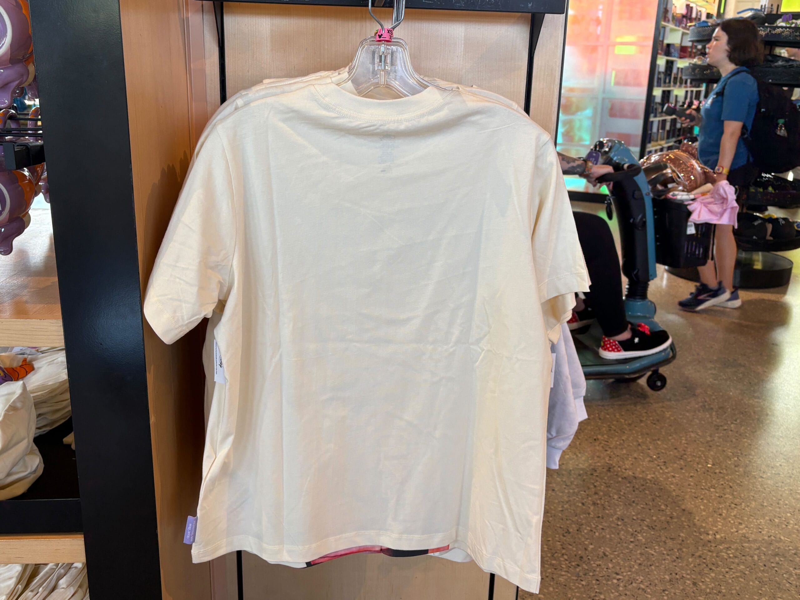 A plain white T-shirt on a hanger displayed in a store, with other shirts and shoppers in the background.