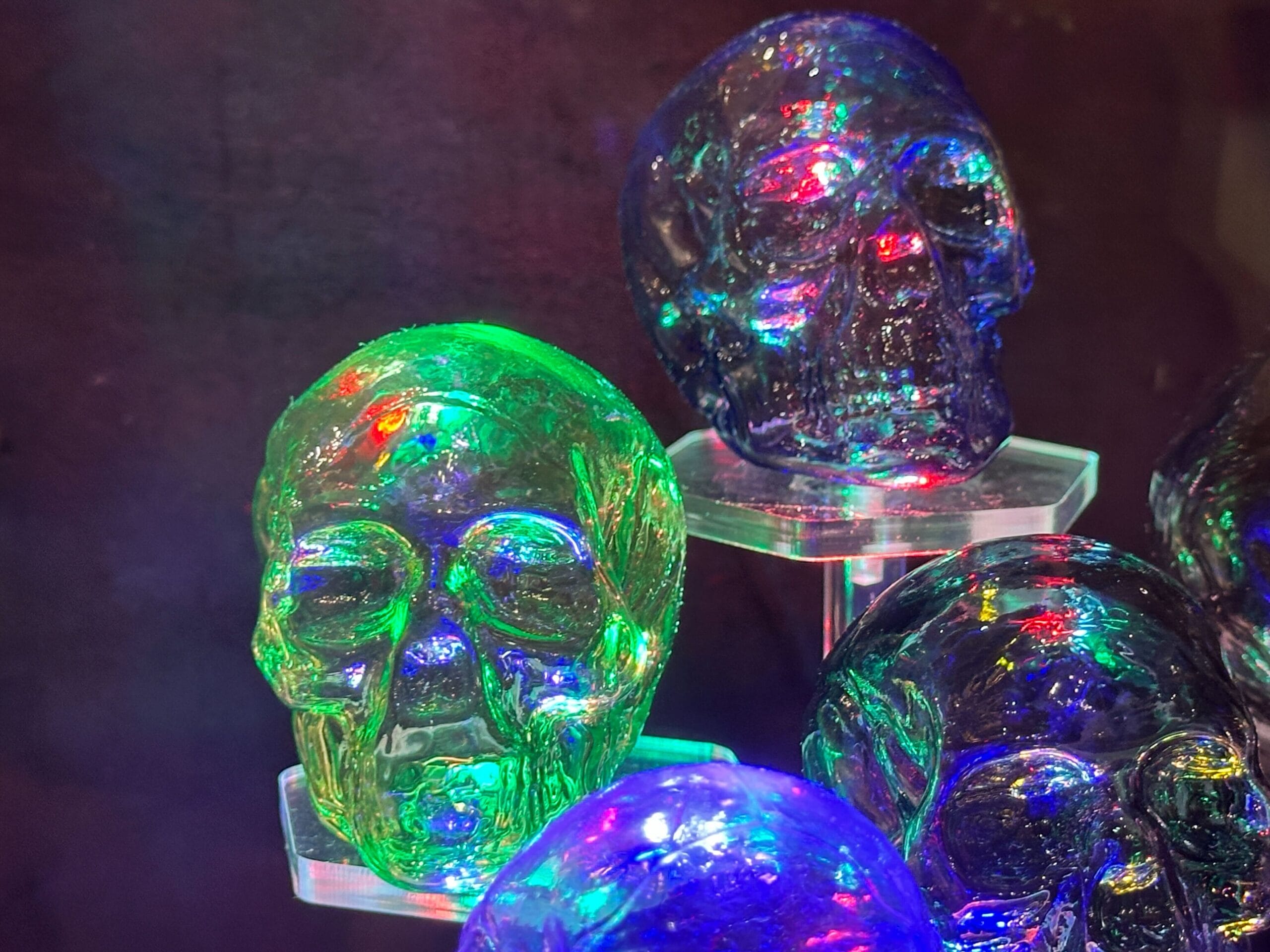 Colorful glass skulls lit with green, blue, and purple hues on clear stands; reminiscent of Disney’s Haunted Mansion decor at night.