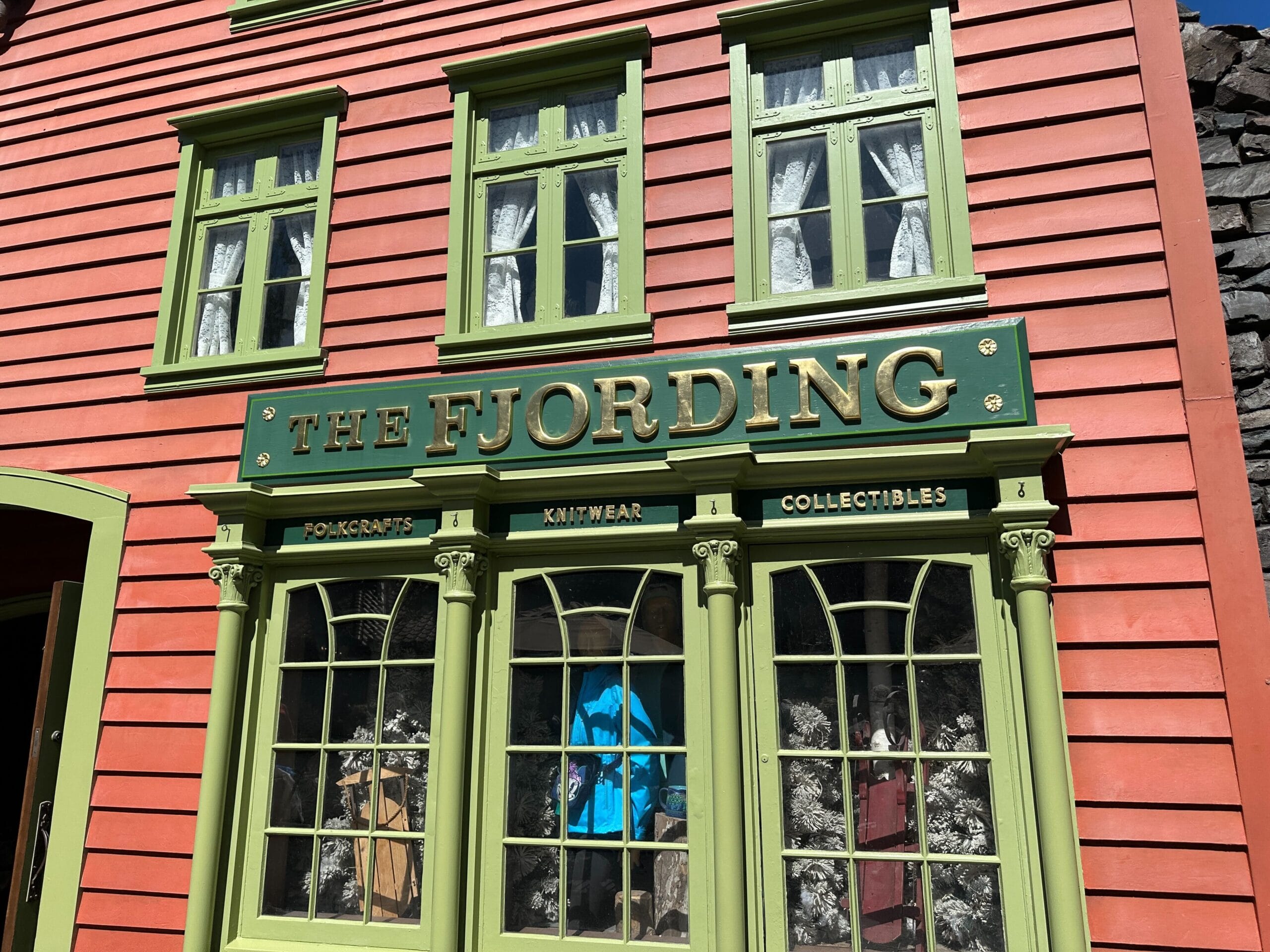 A red and green shopfront labeled "The Fjording" displays folk crafts, knitwear, and collectibles, typical of Epcot’s Norway Pavilion.