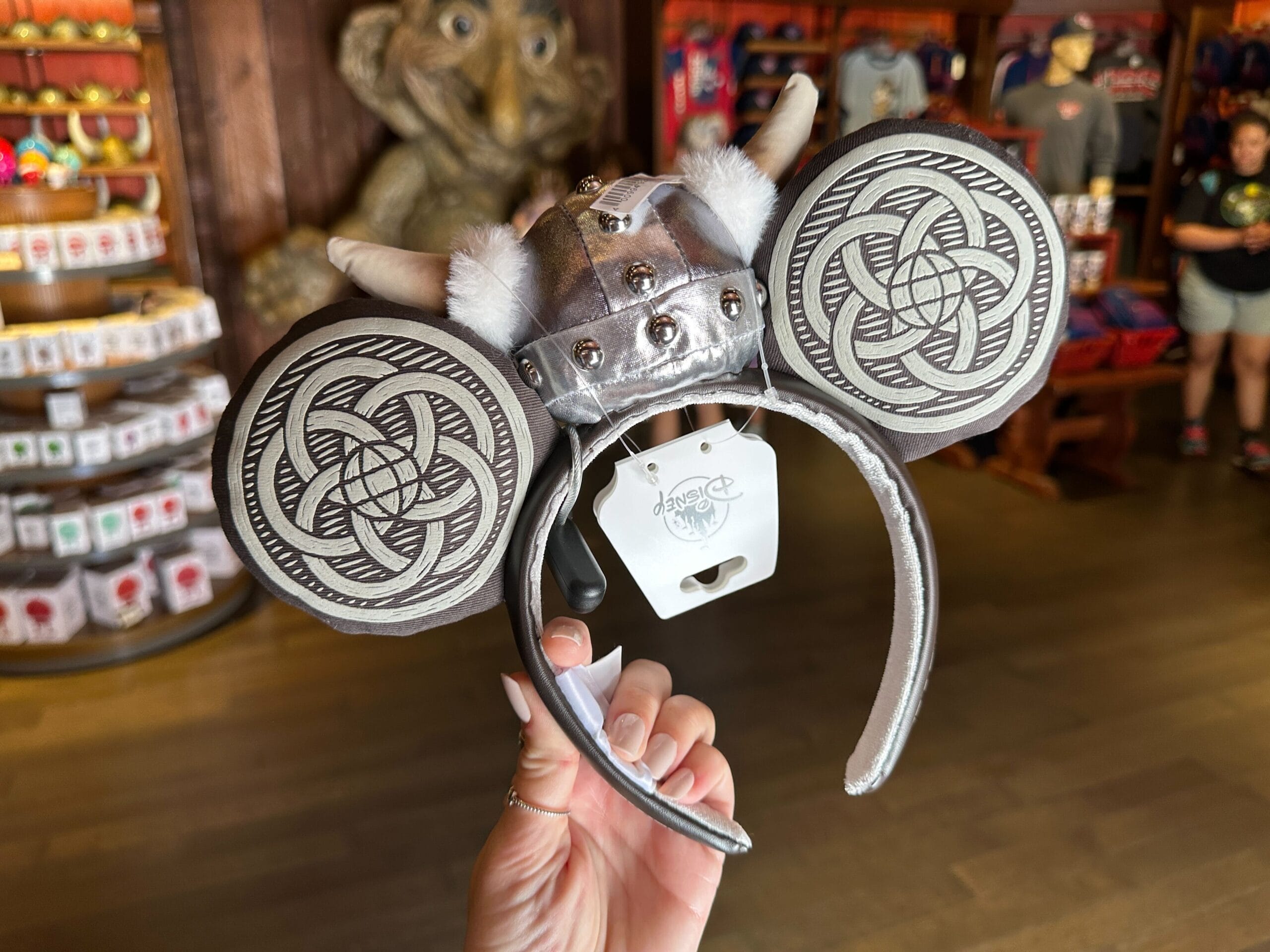 A hand displays silver Viking-themed Mickey Mouse ears inside a Disney park shop, shelves of park merchandise visible behind.