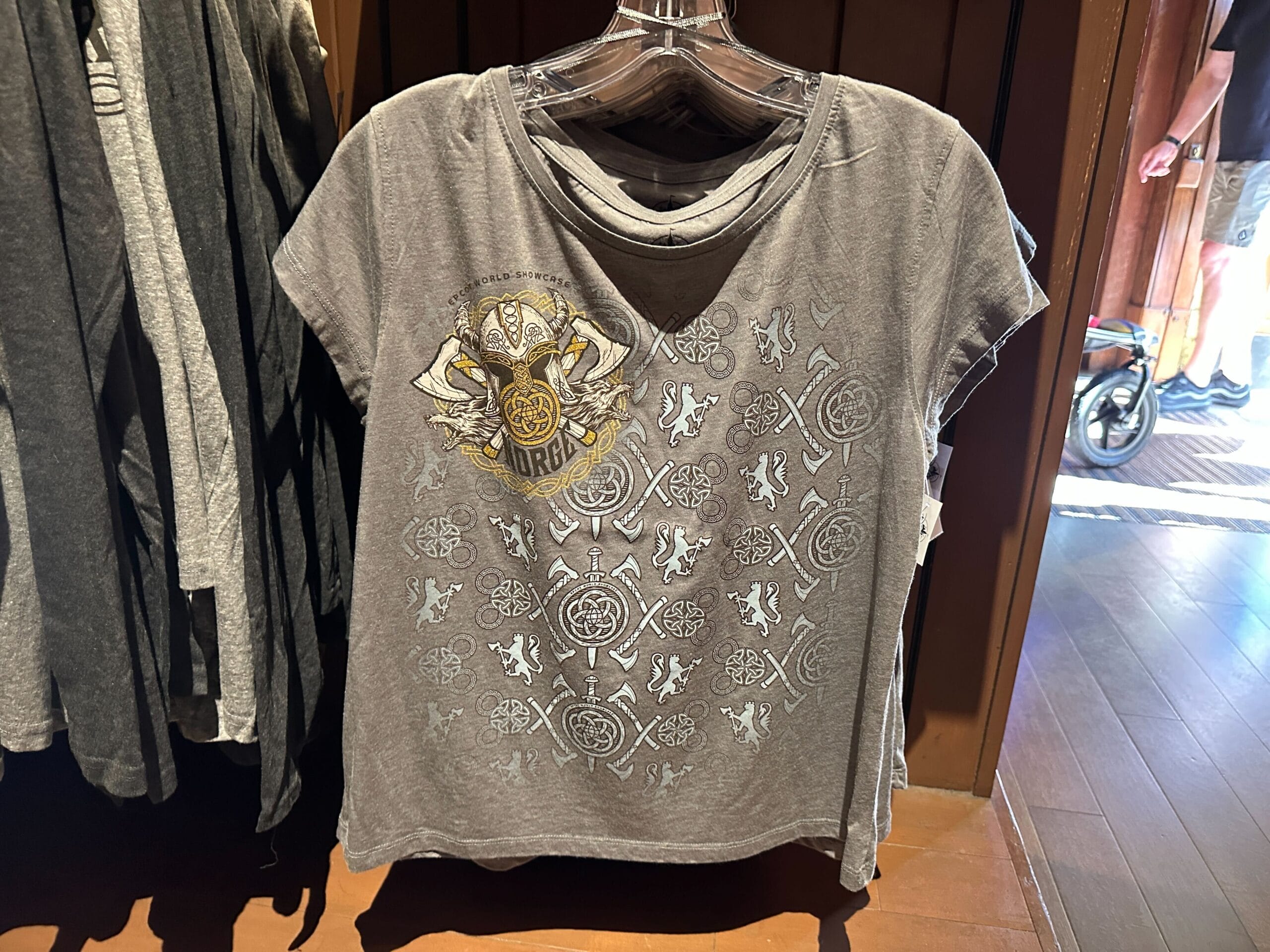 Gray t-shirt with a pattern of Nordic-inspired symbols like axes, shields, and swords, with a larger yellow and gray "Epcot World Showcase Norge" helmet design on the upper right chest.