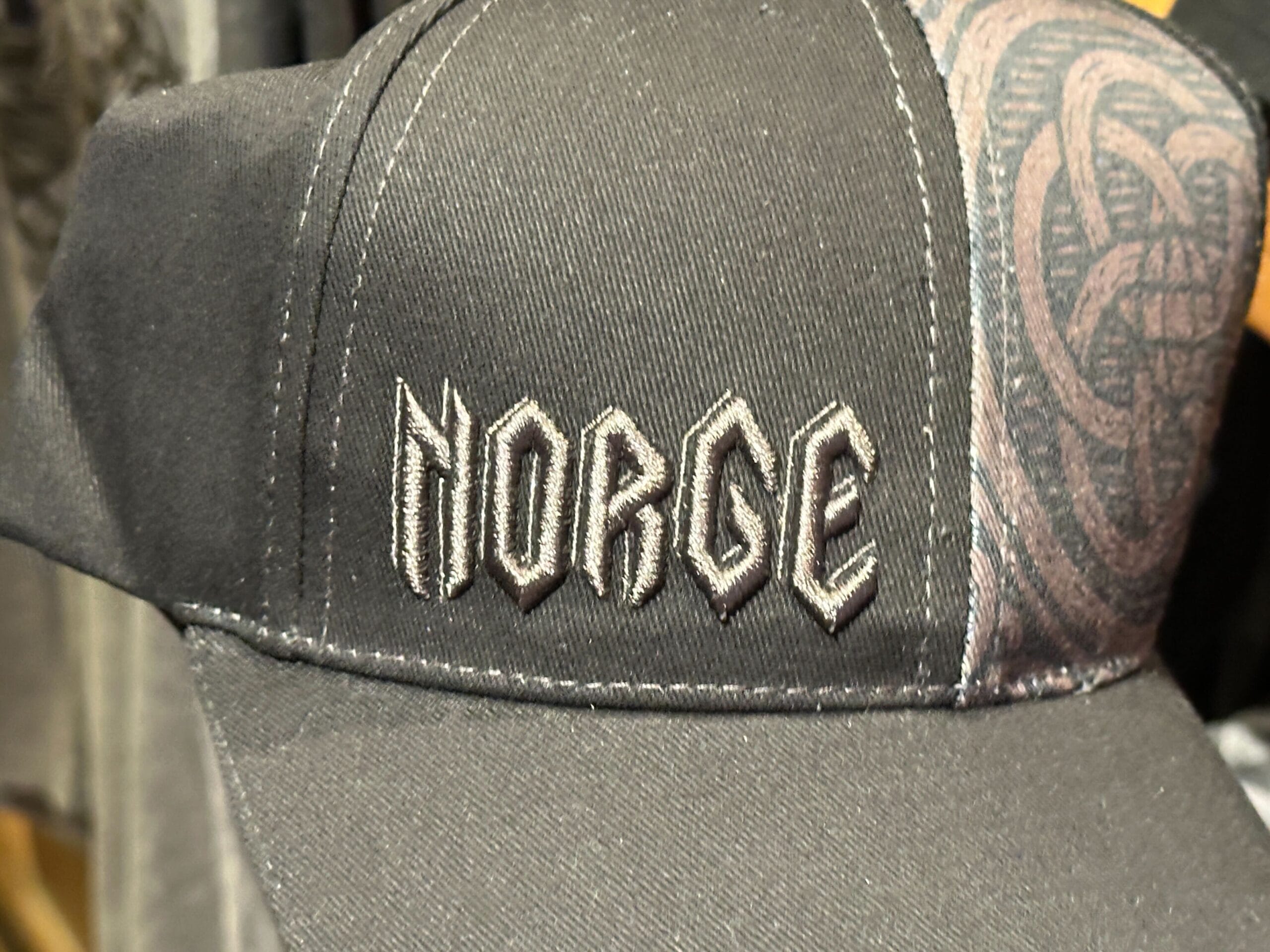 Close up of a black baseball cap with "NORGE" embroidered in a Norse font on the left front panel and an intricate circular Epcot logo-inspired woodcut design on the right.