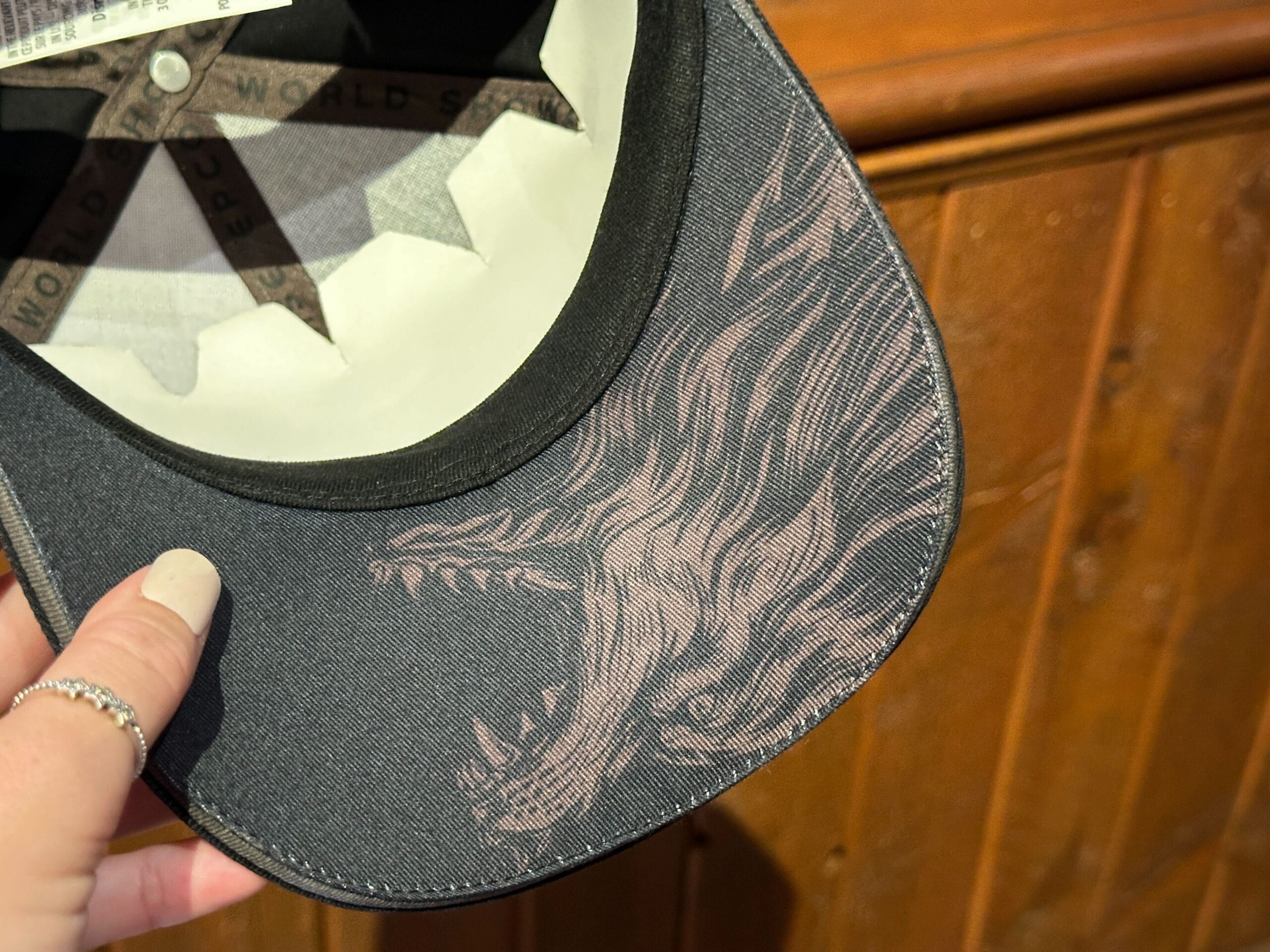 A hand holds a black baseball cap with a wolf underbrim design.