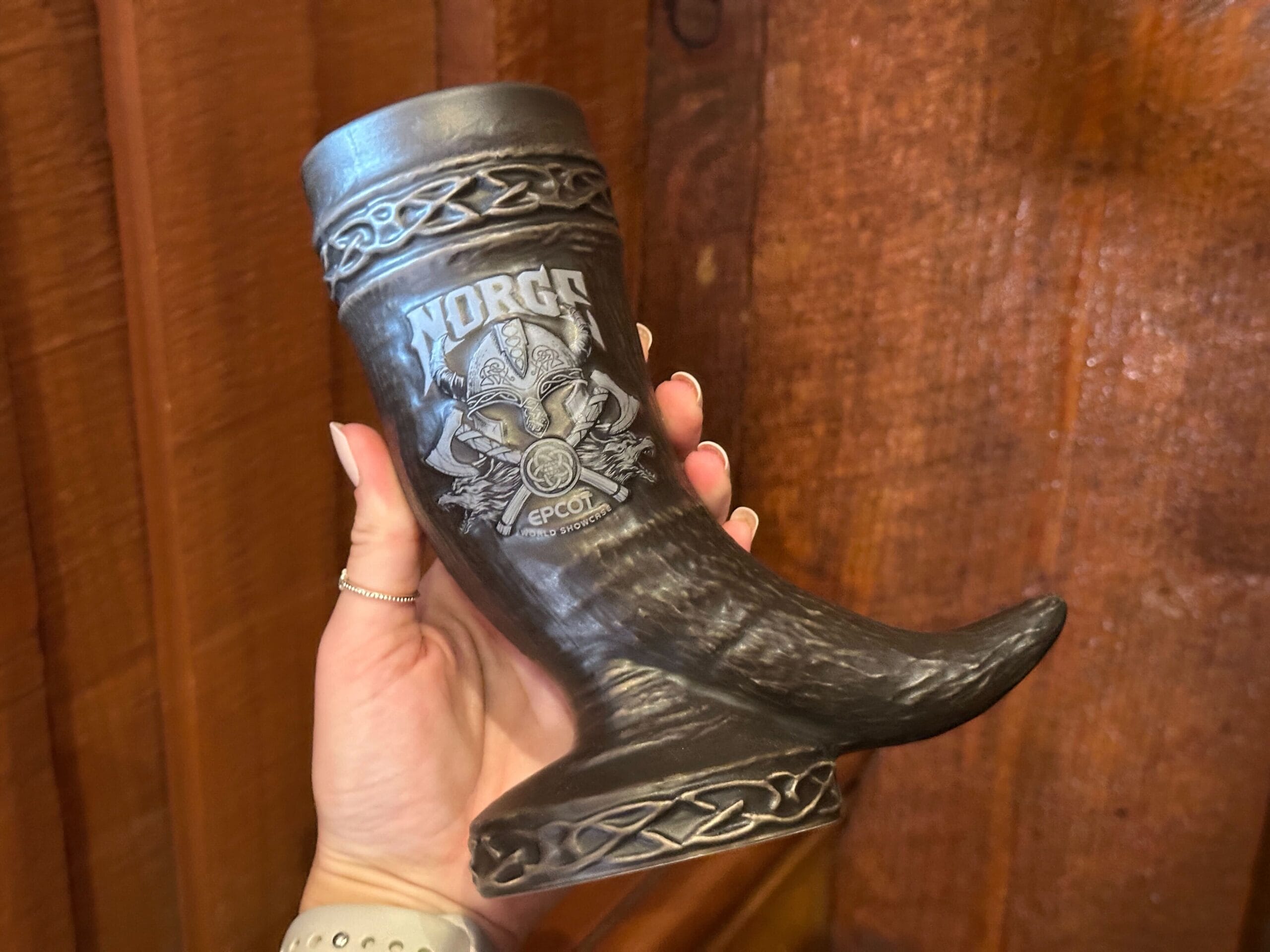 A hand holds a brown ceramic Viking drinking horn-shaped mug engraved with "Norge Epcot World Showcase" and a Viking helmet design. A knotwork pattern is sculpted around the top rim. The "horn" rests on an attached round base with a knotwork pattern.