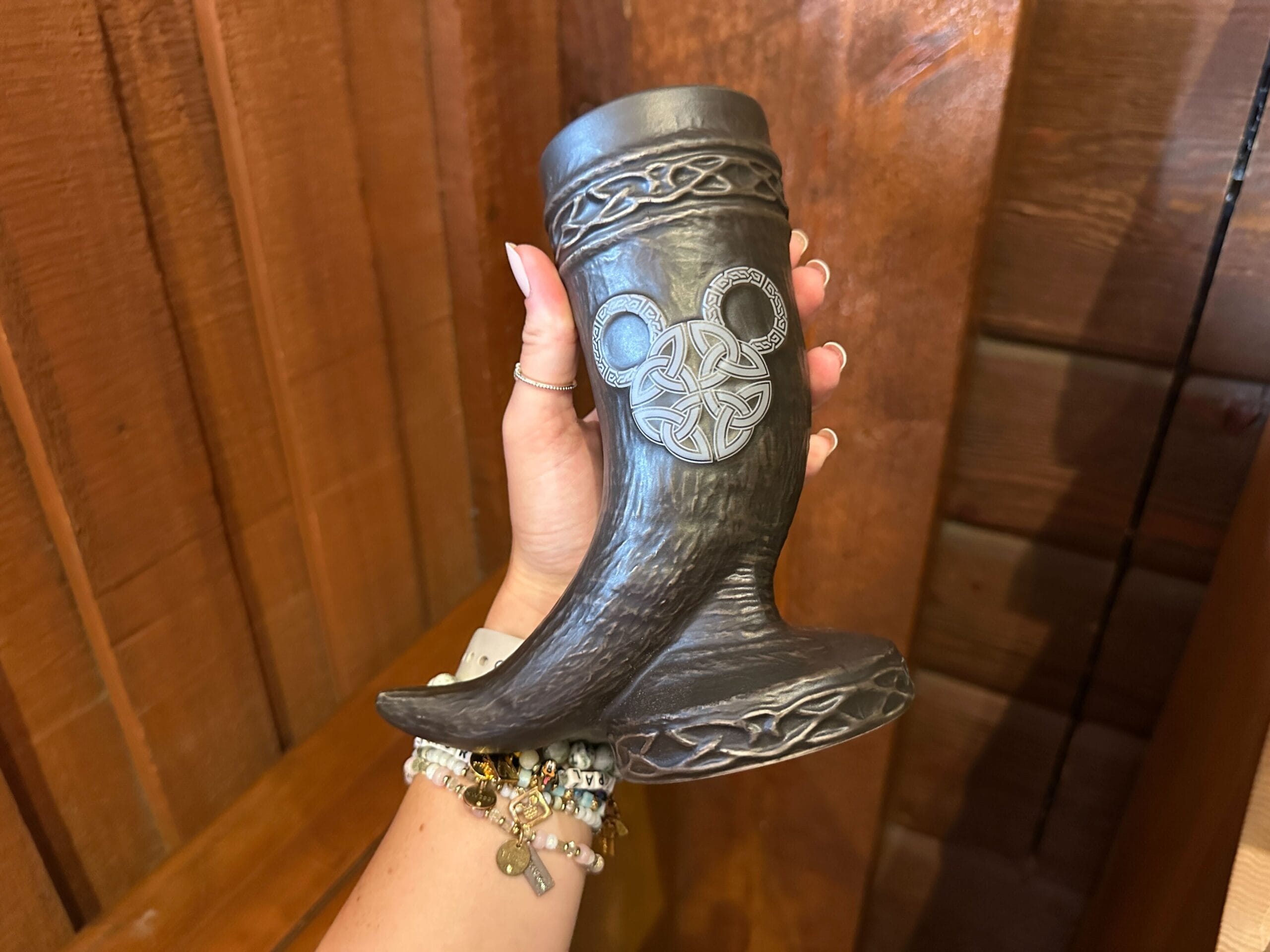 A hand holds a brown ceramic Viking drinking horn-shaped mug with a Nordic-inspired Mickey head design. A knotwork pattern is sculpted around the top rim. The "horn" rests on an attached round base with a knotwork pattern.