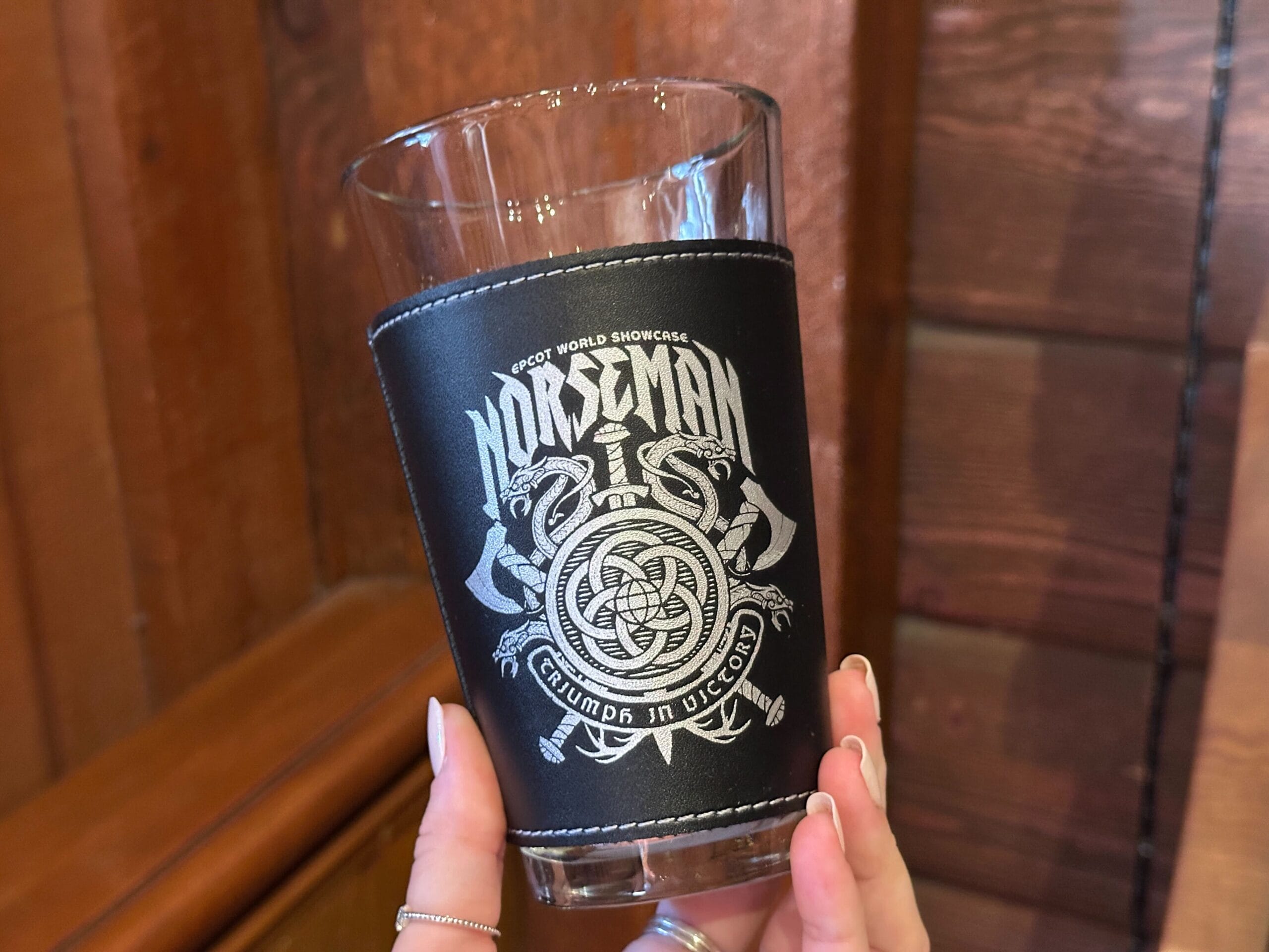 A hand holds a clear pint glass with a black pleather sleeve with white stitching and an "Epcot World Showcase Norseman" graphic featuring a Viking shield, axes, and dragons.