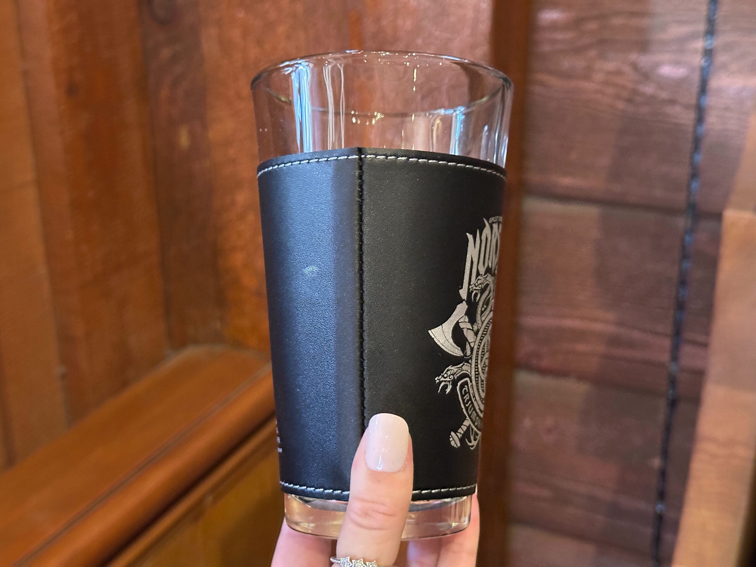 A hand holds a clear pint glass with a black pleather sleeve with white stitching, showing a vertical seam. A white Nordic design is visible on the left.