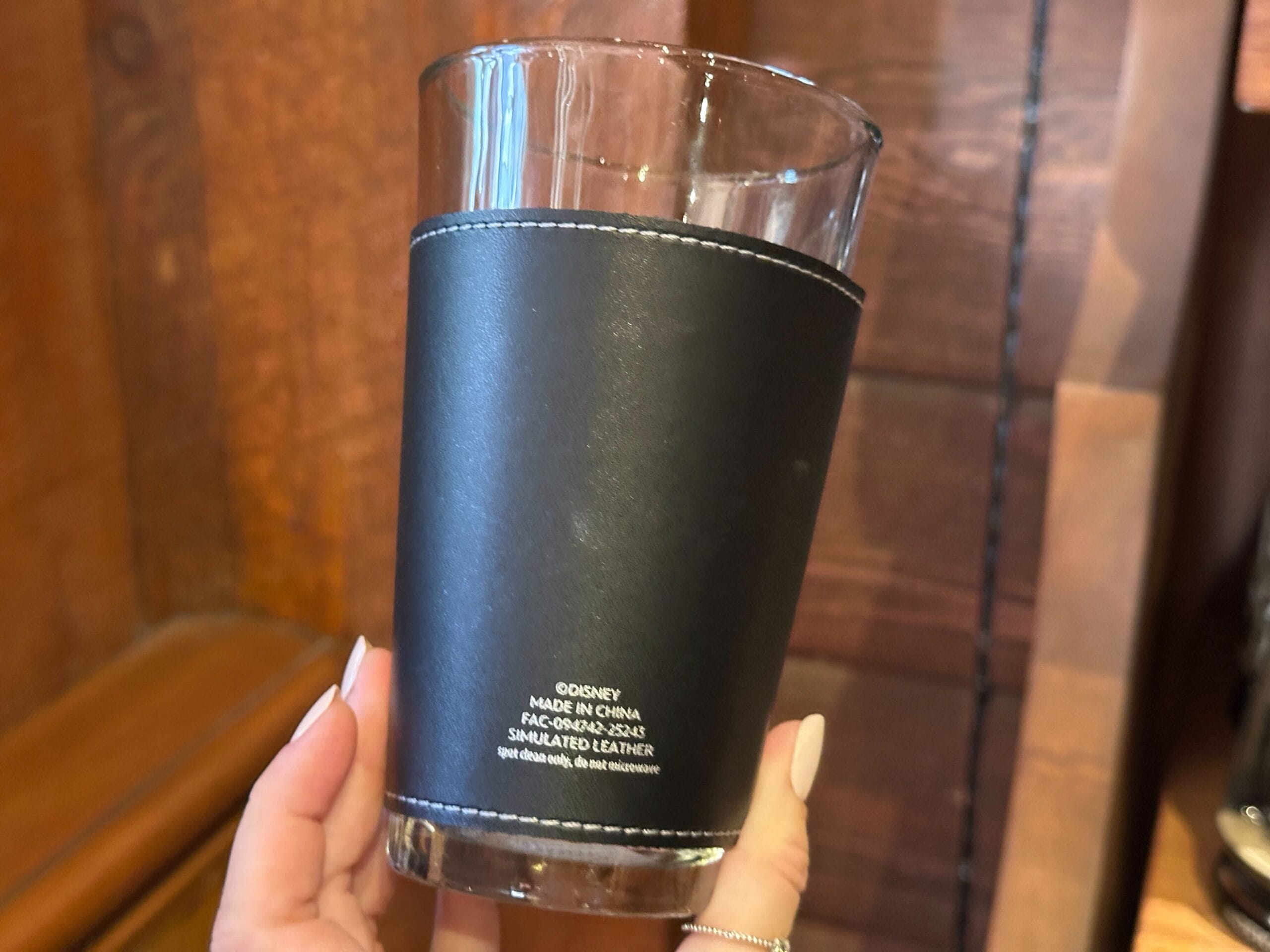 A hand holds a clear pint glass with a black pleather sleeve with white stitching. Manufacturer information is printed in small white text near the bottom.