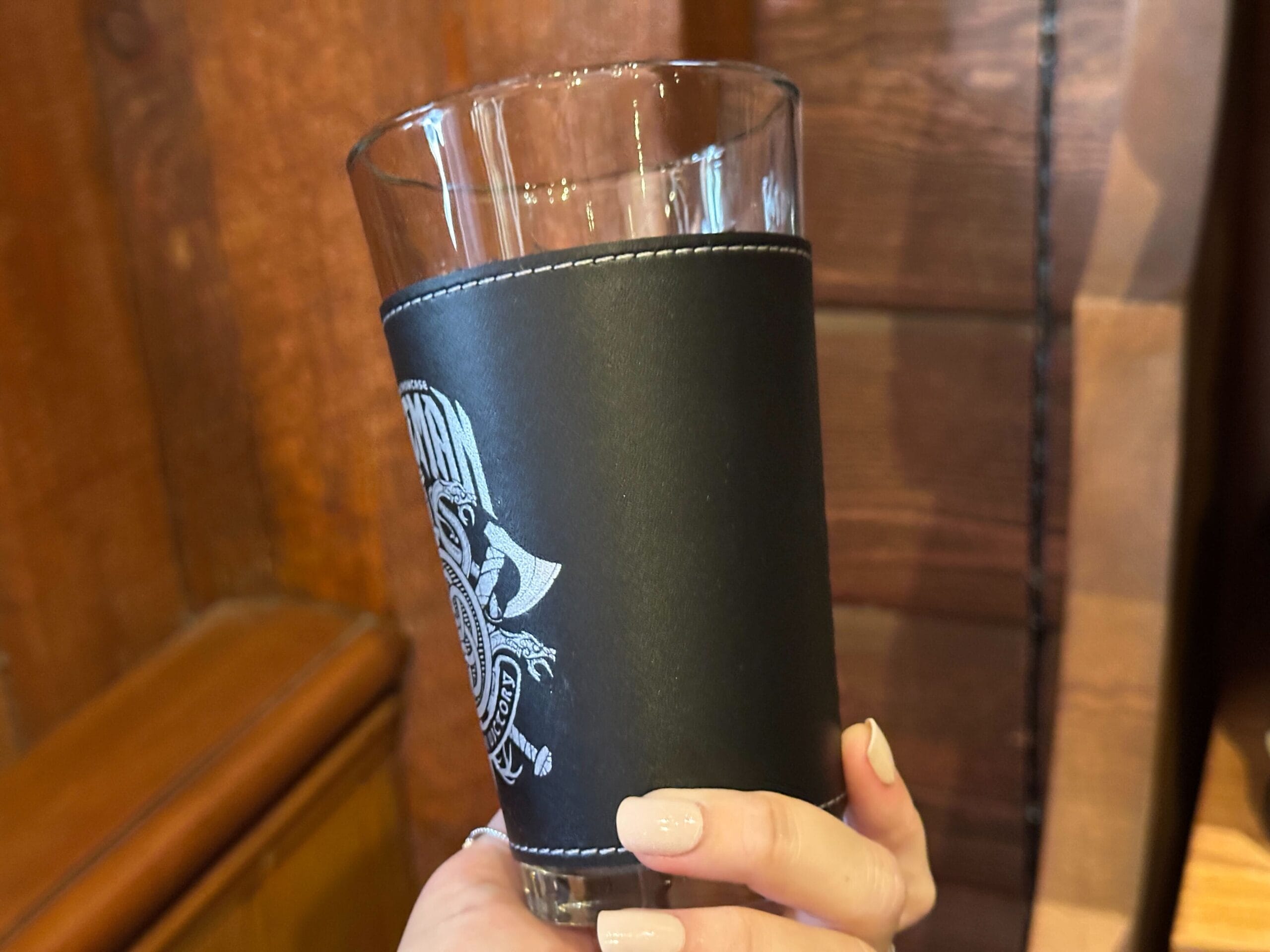 A hand holds a clear pint glass with a black pleather sleeve with white stitching. A white Nordic design is visible on the right.
