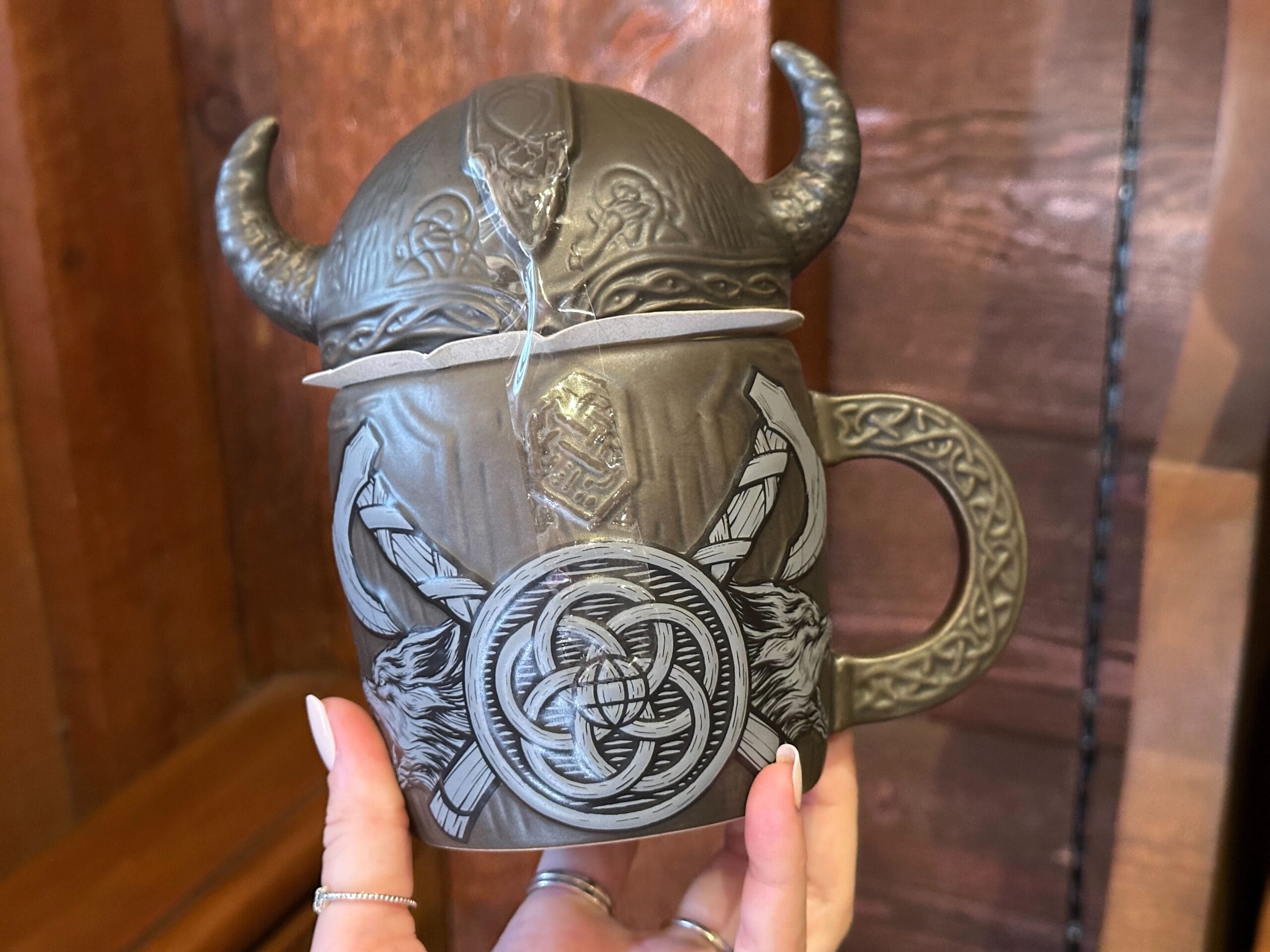 A hand holds a brown and gray Viking-inspired mug with featured a horned helmet lid and a Nordic design of axes and wolves around a shield. The gray handle has a sculpted knotwork pattern.