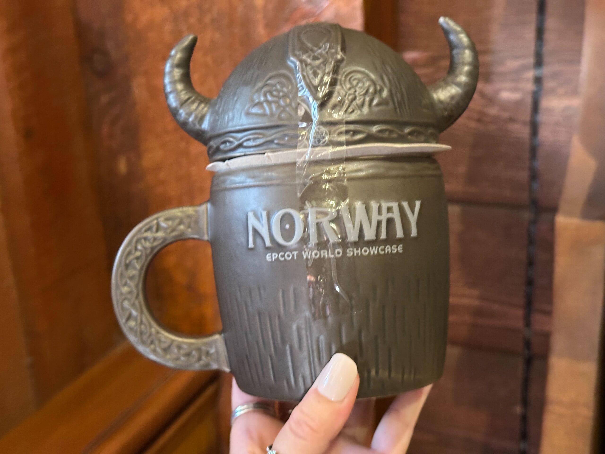 A hand holds a brown and gray Viking helmet mug reading "Norway Epcot World Showcase." The gray handle has a molded knotwork pattern.