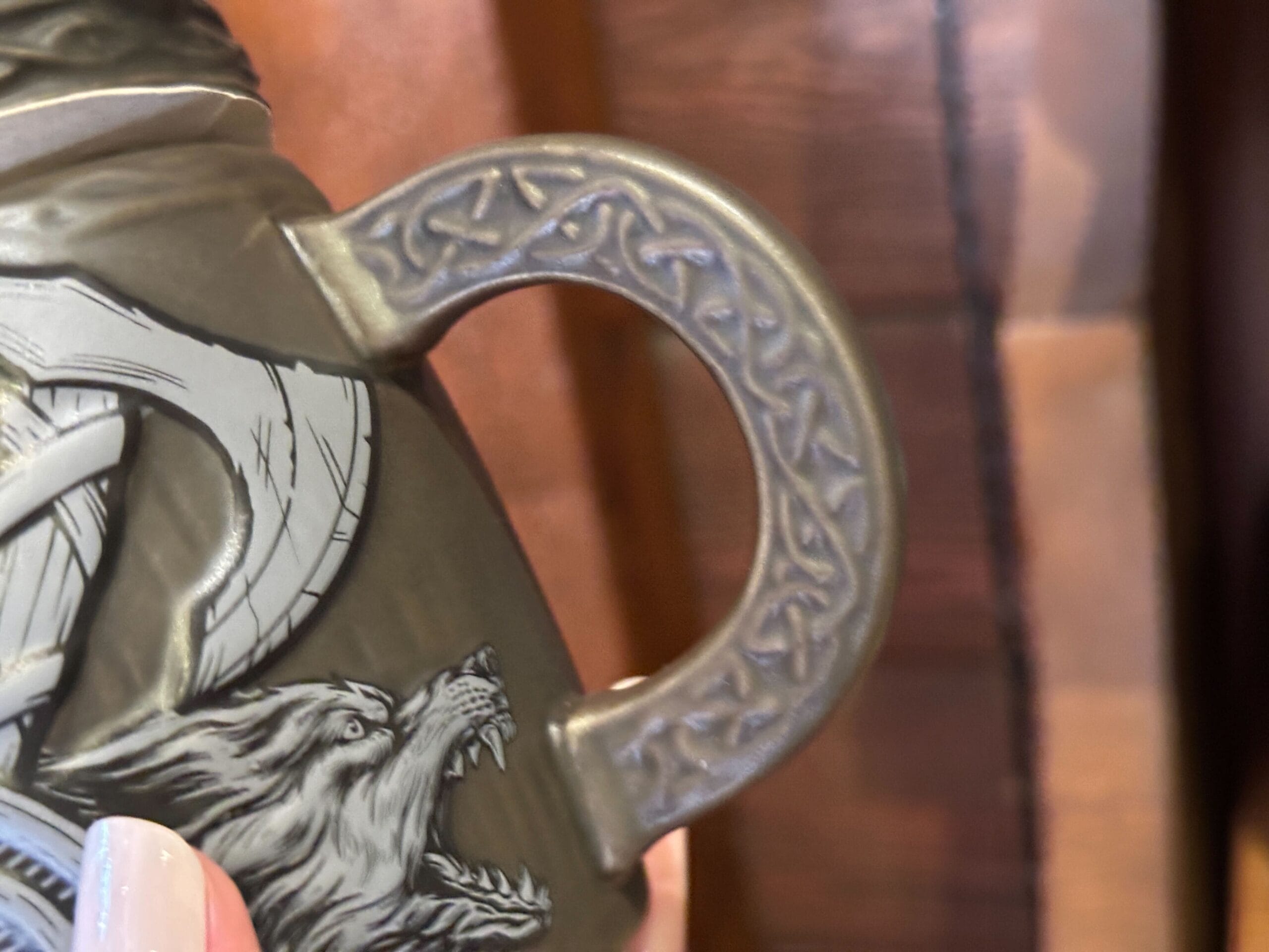 A close-up of a gray mug handle with sculpted patterns; on the brown mug body next to it is a snarling wolf below an axe head.
