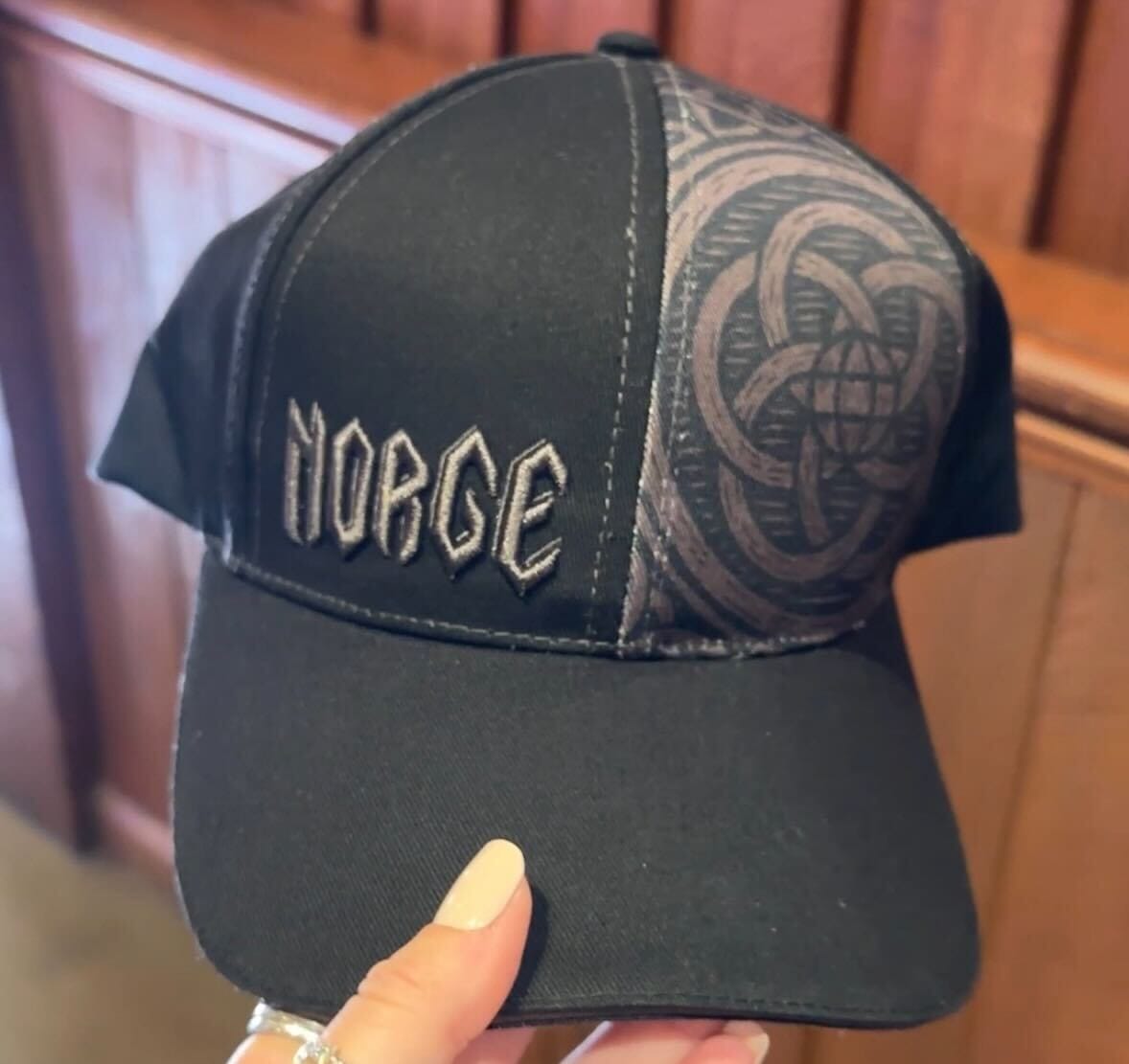 A hand holds a black baseball cap with "NORGE" embroidered in a Norse font on the left front panel and an intricate circular Epcot logo-inspired woodcut design on the right.