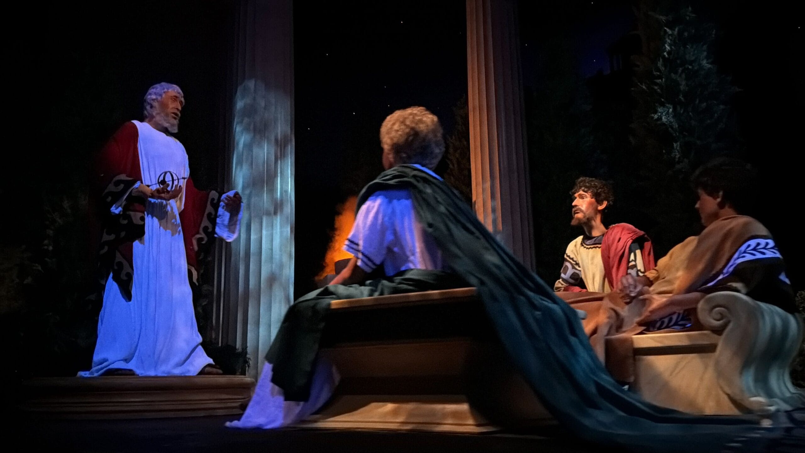 Three men in ancient robes listen to a speaker between pillars at night, evoking Spaceship Earth at EPCOT with its iconic ambiance.