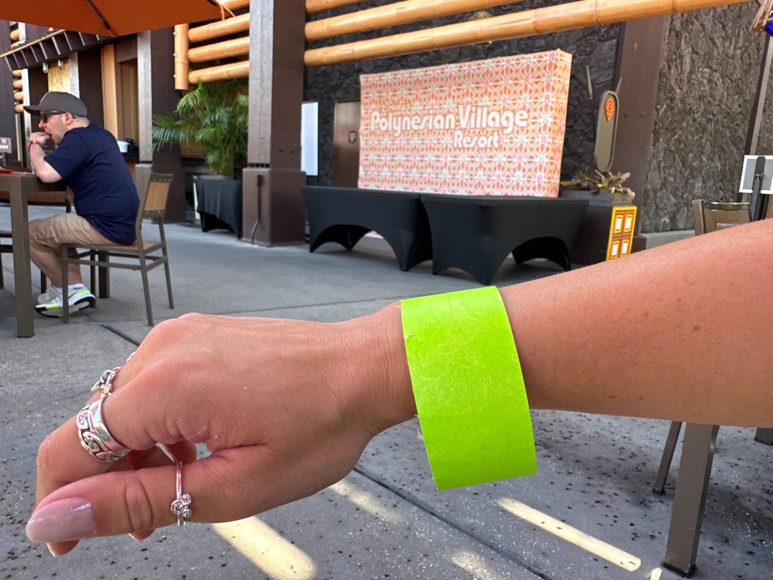 A hand with a neon green wristband holds a Green Tiki Mug in front of the Disney Polynesian Village Resort sign, no castle visible.