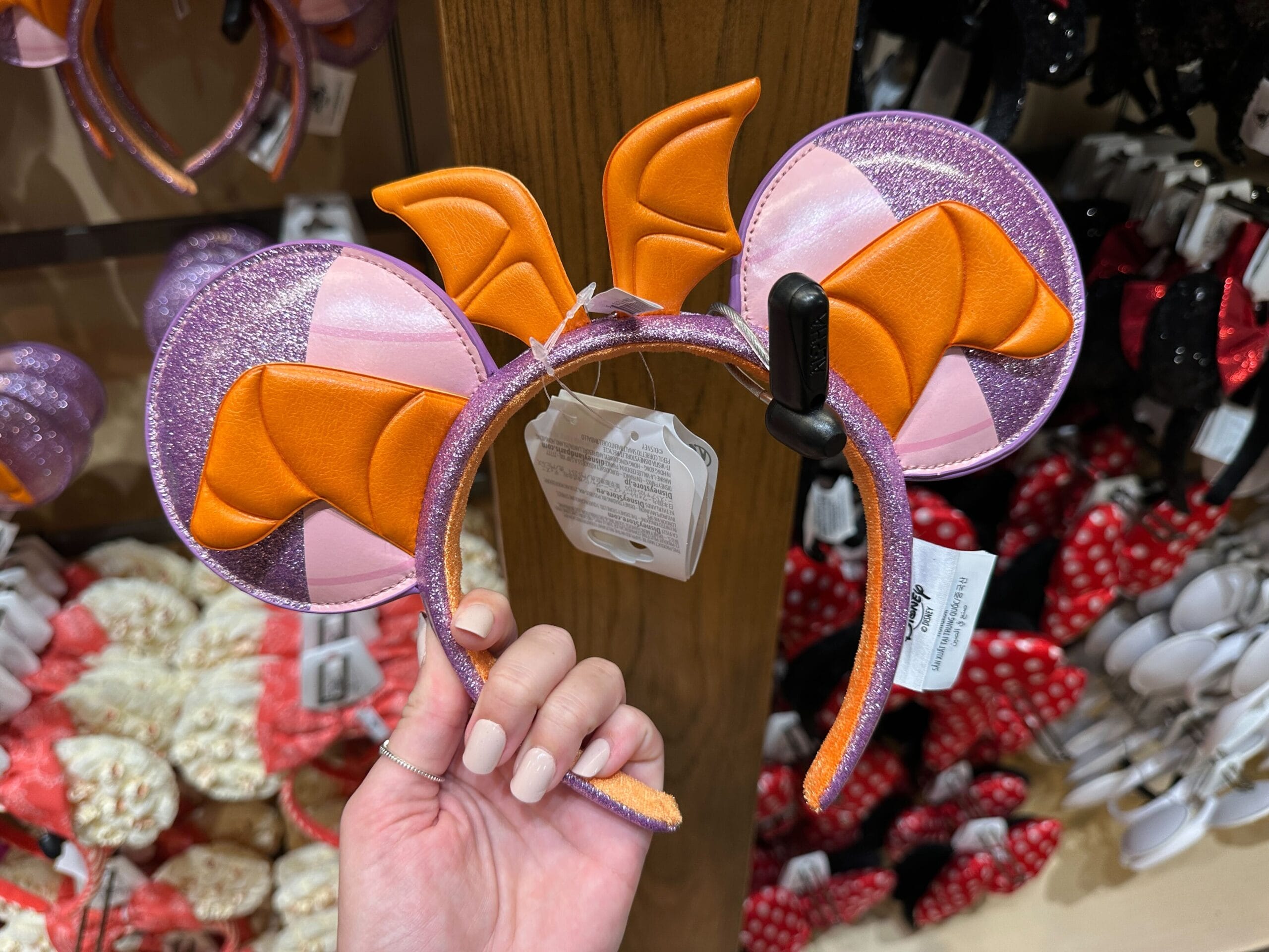 A hand holds a headband with orange dragon wings and purple glittery ears, shown in a Disney park shop among other whimsical headbands.