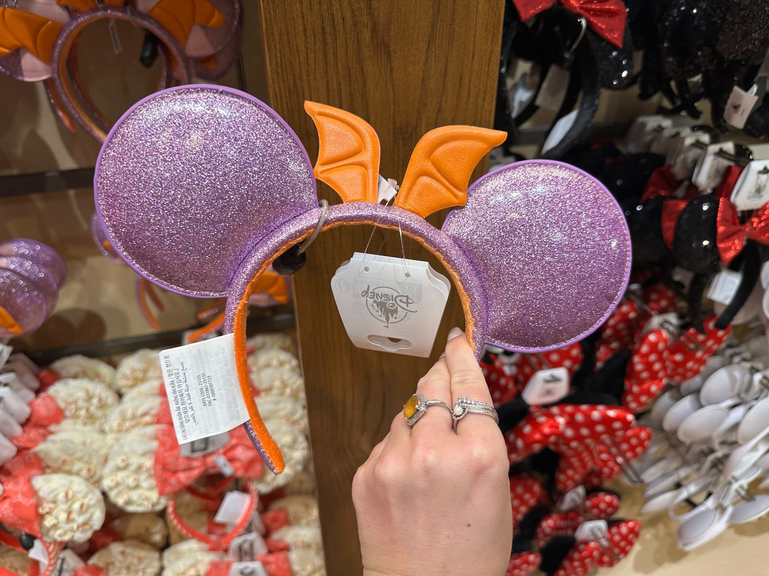 A hand displays a sparkly purple Mickey ear headband with orange bat wings, spotted in a Disney park shop with magical merchandise.