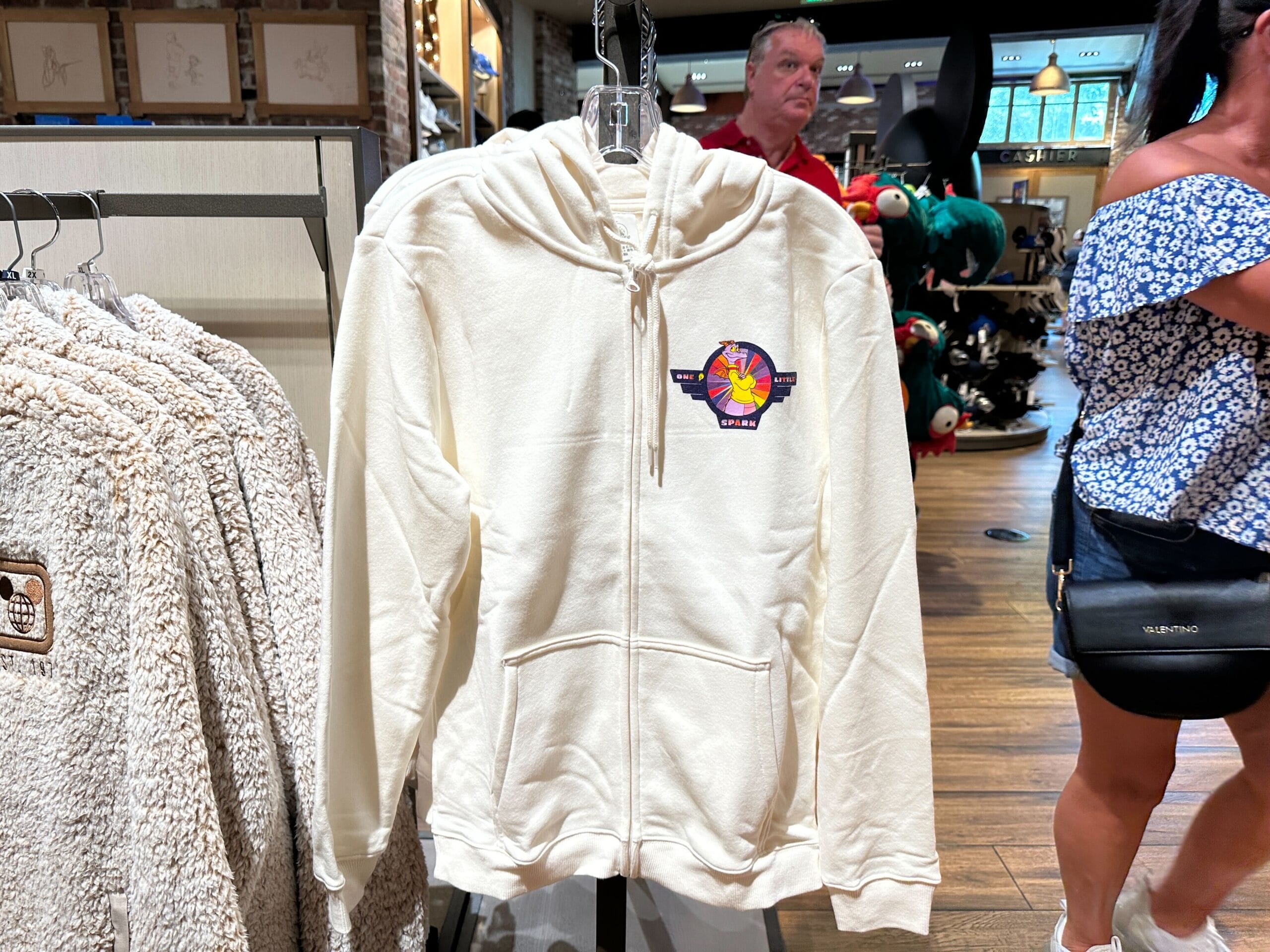 A white zip-up hoodie featuring Disney’s vibrant Space Mountain logo across the chest, displayed on a rack in a Disney park shop.