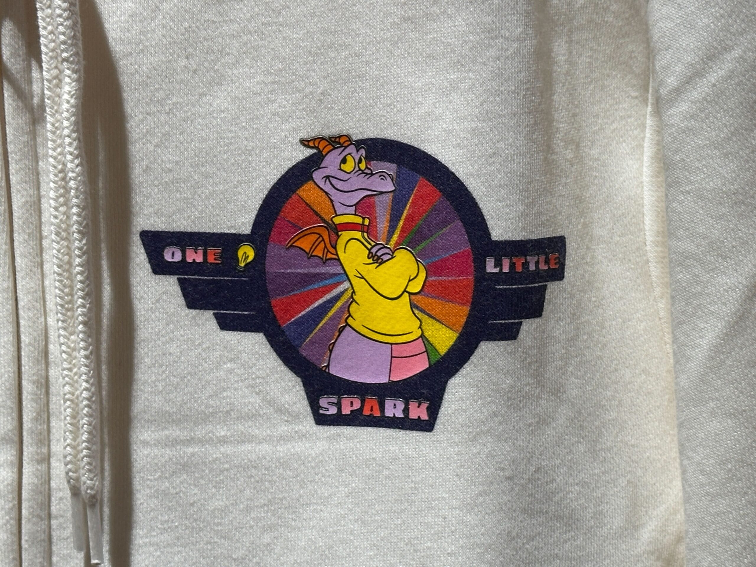 Figment the purple dragon in a yellow shirt with "One Little Spark" text, set on a vibrant circular Disney-themed background.