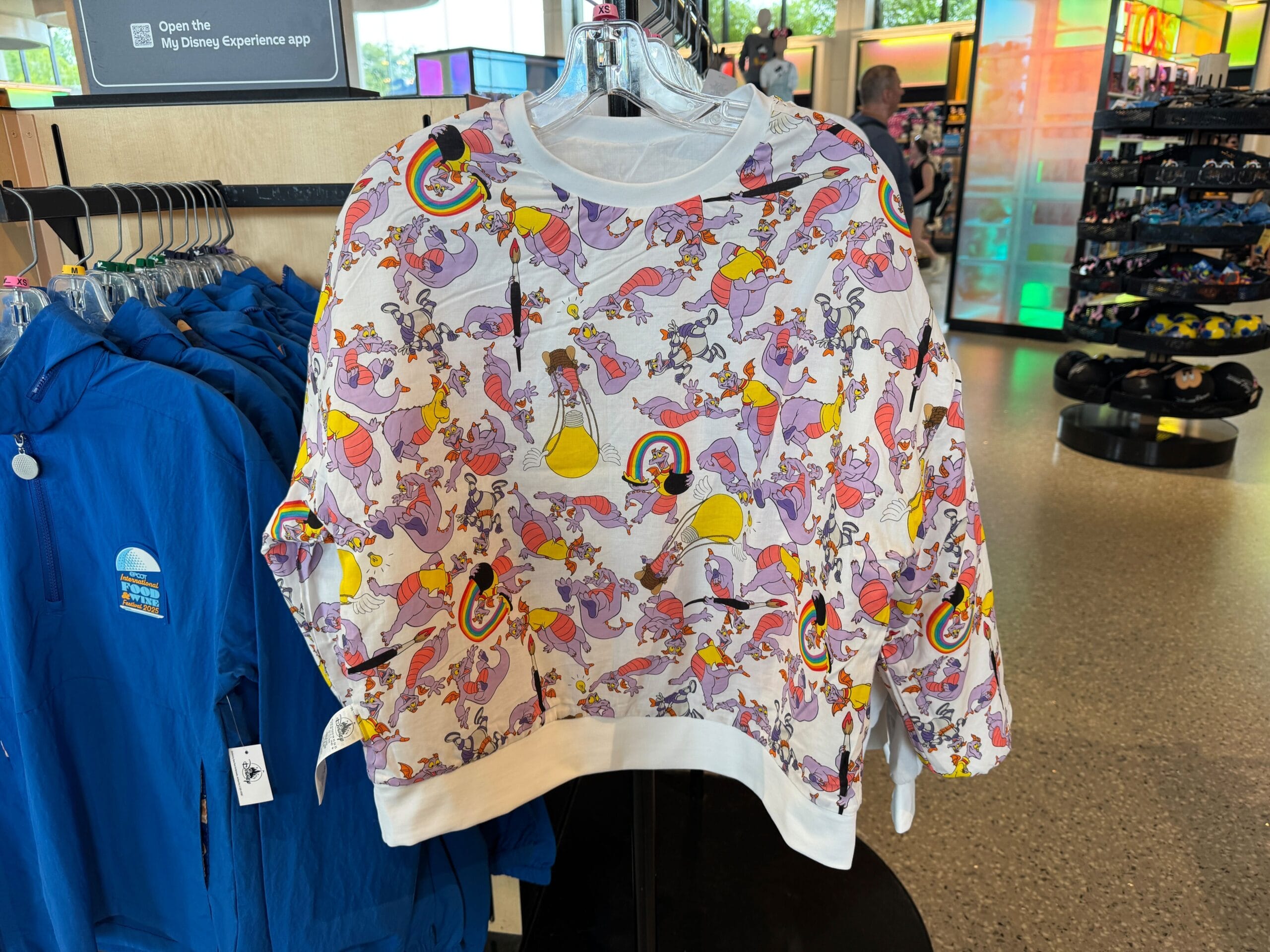 White sweatshirt with colorful cartoon character and lab equipment print hanging in a store next to blue jackets.