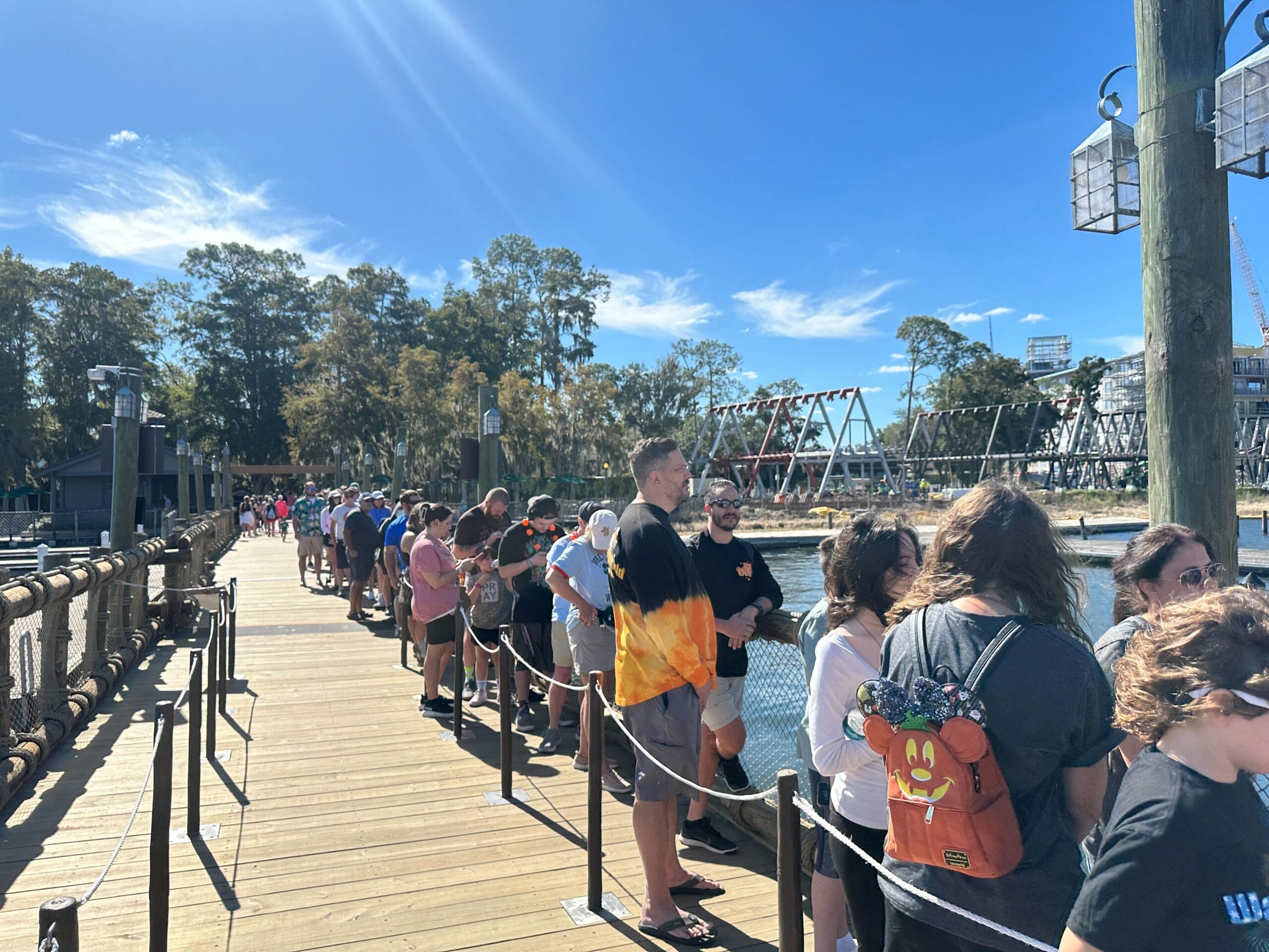 Guests wait in a long queue on a wooden dock by the water at a Disney park, with attractions and trees visible in the background.