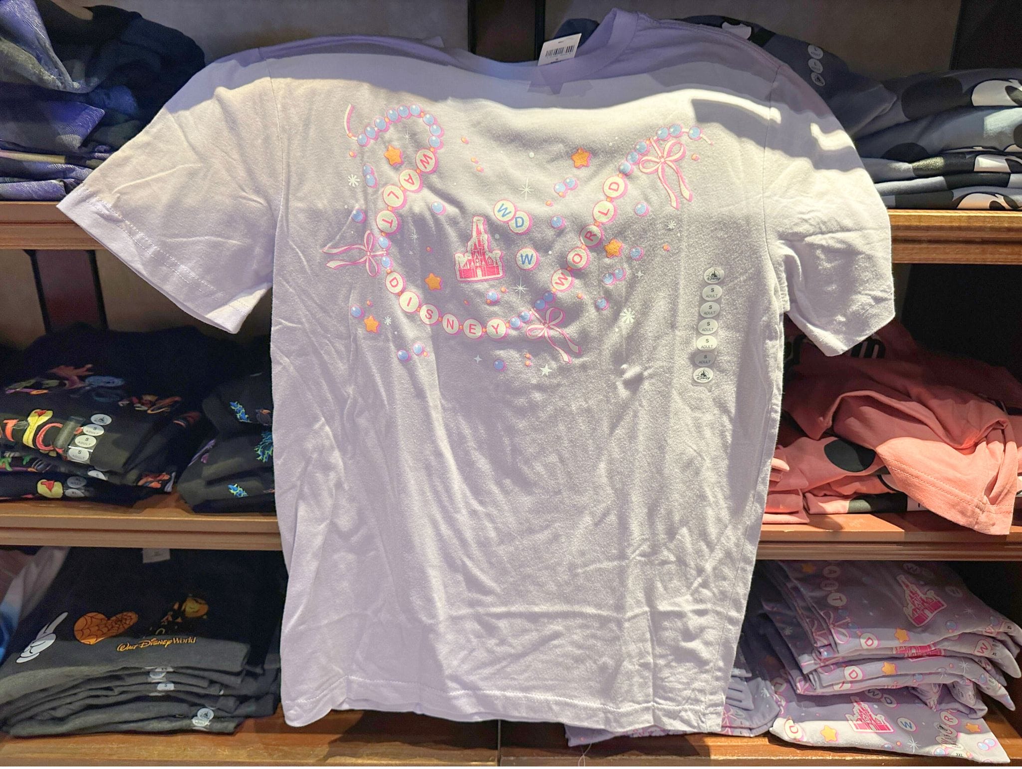 White T-shirt with pastel castle, bows, and alphabet beads design, displayed on a shelf with folded shirts.