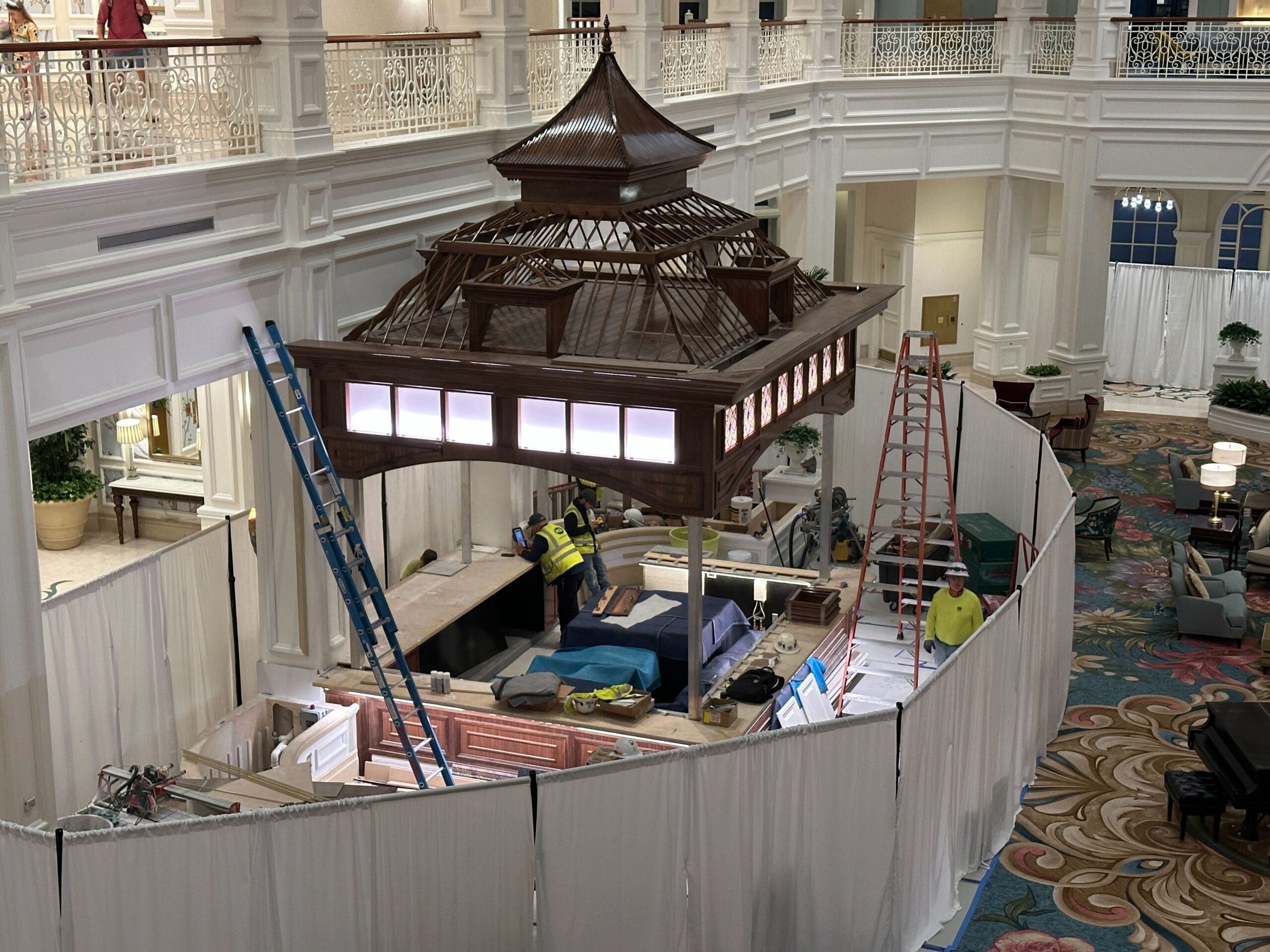 Workers refurbish an indoor space at a Disney park, building a wooden pavilion surrounded by ladders and white curtains.